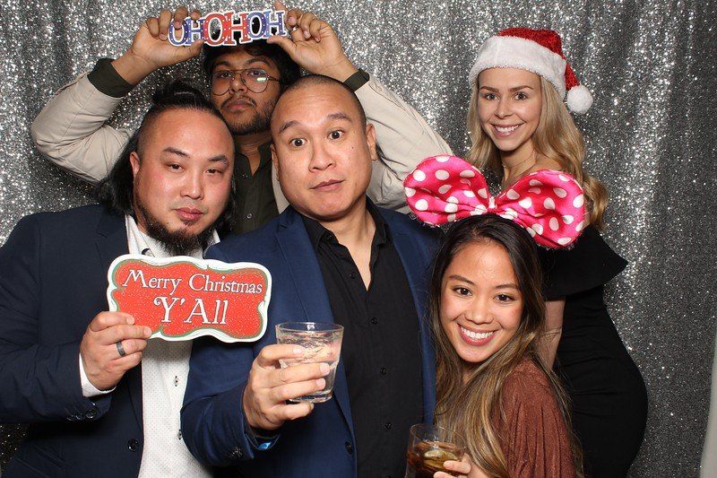 Group of six people at a party in front of a silver backdrop, smiling, holding props, and dressed up for the holidays.