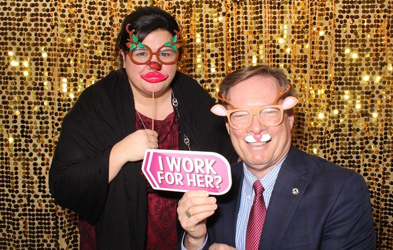 Two people pose with photo booth props, gold backdrop. Woman wears reindeer glasses, holding 