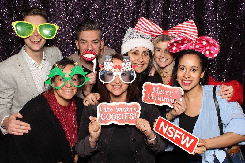 Group of smiling people in a photo booth wearing novelty glasses and holding holiday-themed props.