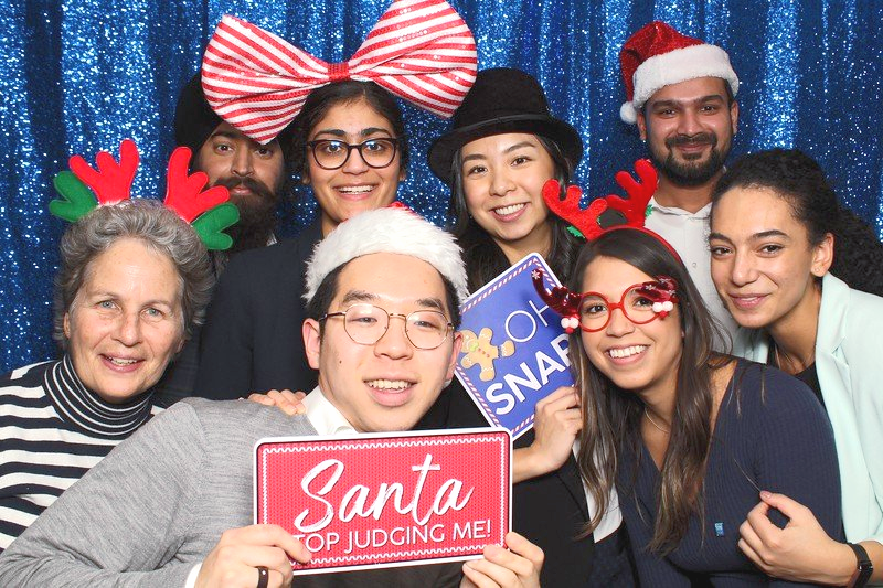 Group of people smiling at a photo booth. Holding props such as Santa hats, reindeer antlers, and signs. Blue backdrop.