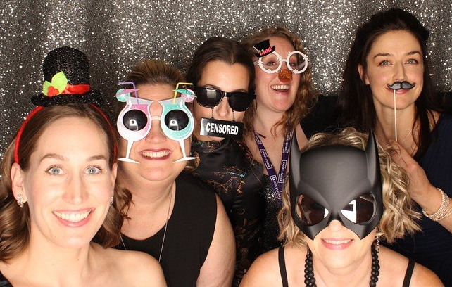 Corporate holiday event photo booth with a group of people posing with props against a silver shiny backdrop.