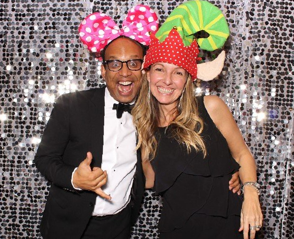 Man and woman smiling in photo booth, wearing novelty hats, in front of a silver sequin backdrop.