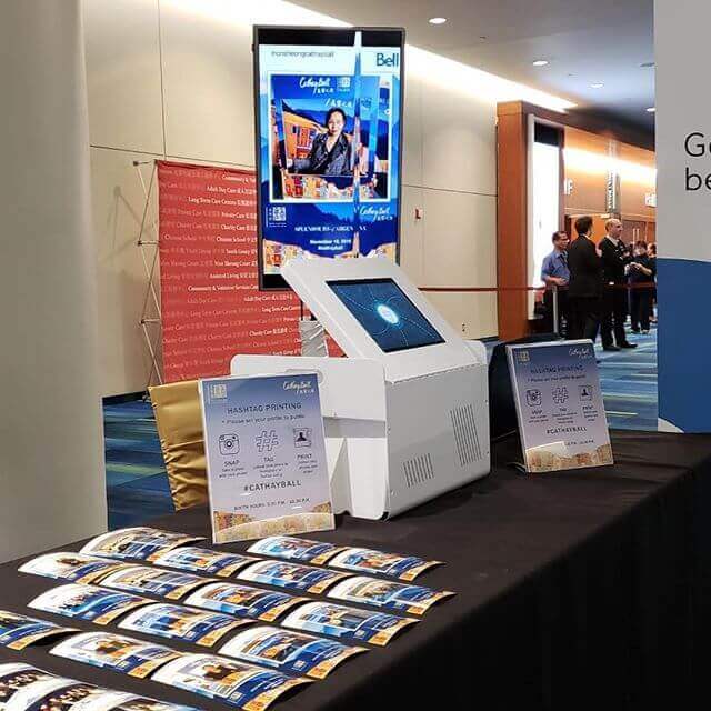 An information booth with brochures, digital display, and touchscreen kiosk. People are visible in the background.
