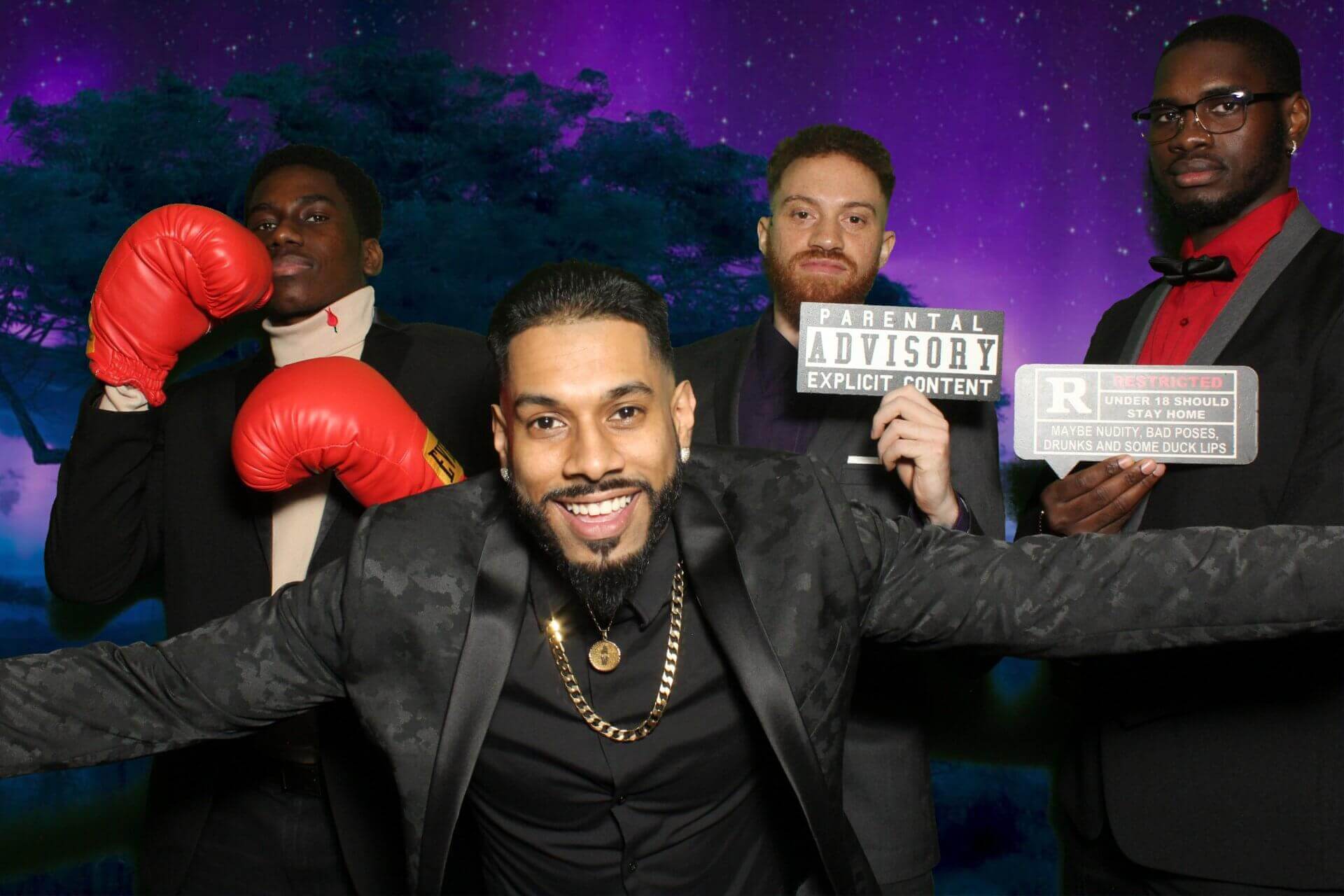 Four people pose in a photo booth. One wears boxing gloves, the others hold parental advisory and R-rated signs. Purple backdrop.