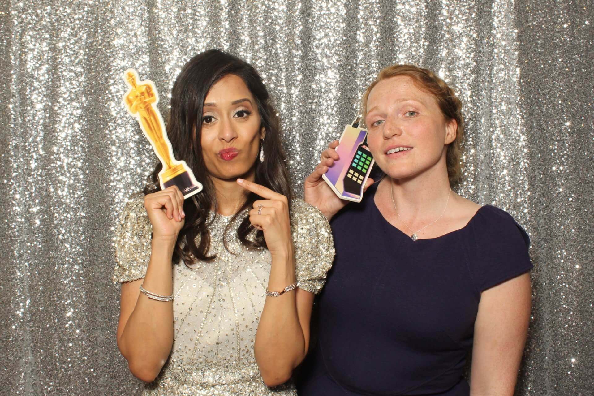 Two women pose with photo booth props in front of a sparkly background. One points to an Oscar prop, the other holds a phone prop.