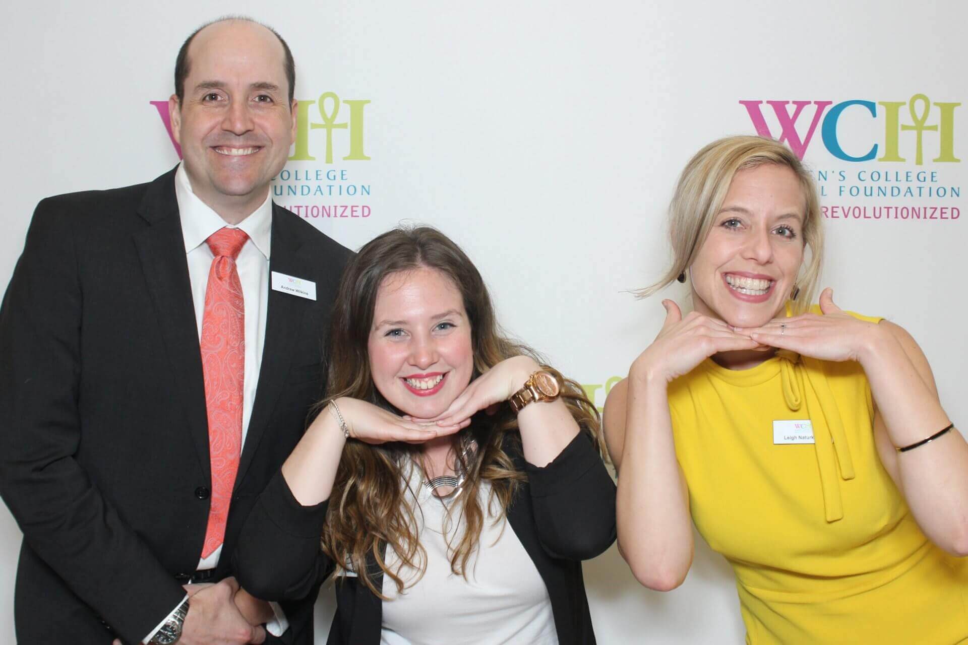 Three people posing in front of a backdrop with a logo; two women with their hands on their chins, man in a suit smiles.