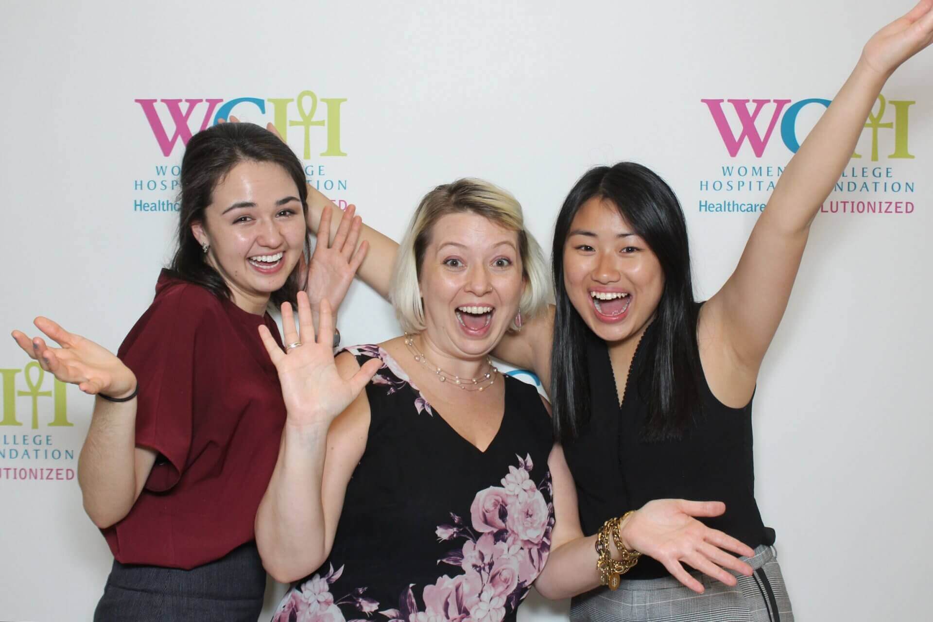 Three women smiling and posing in front of a backdrop with a logo.