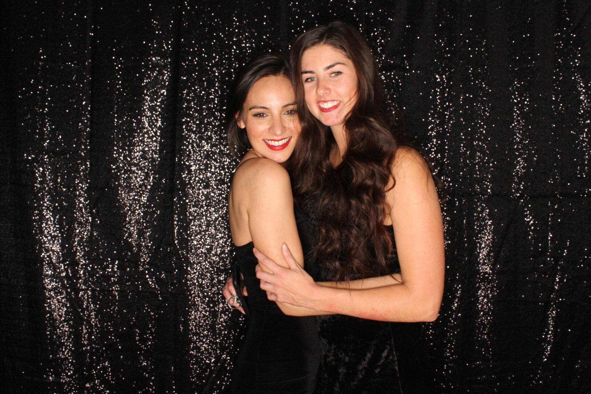 Two women smiling, hugging in front of a sequined black backdrop.