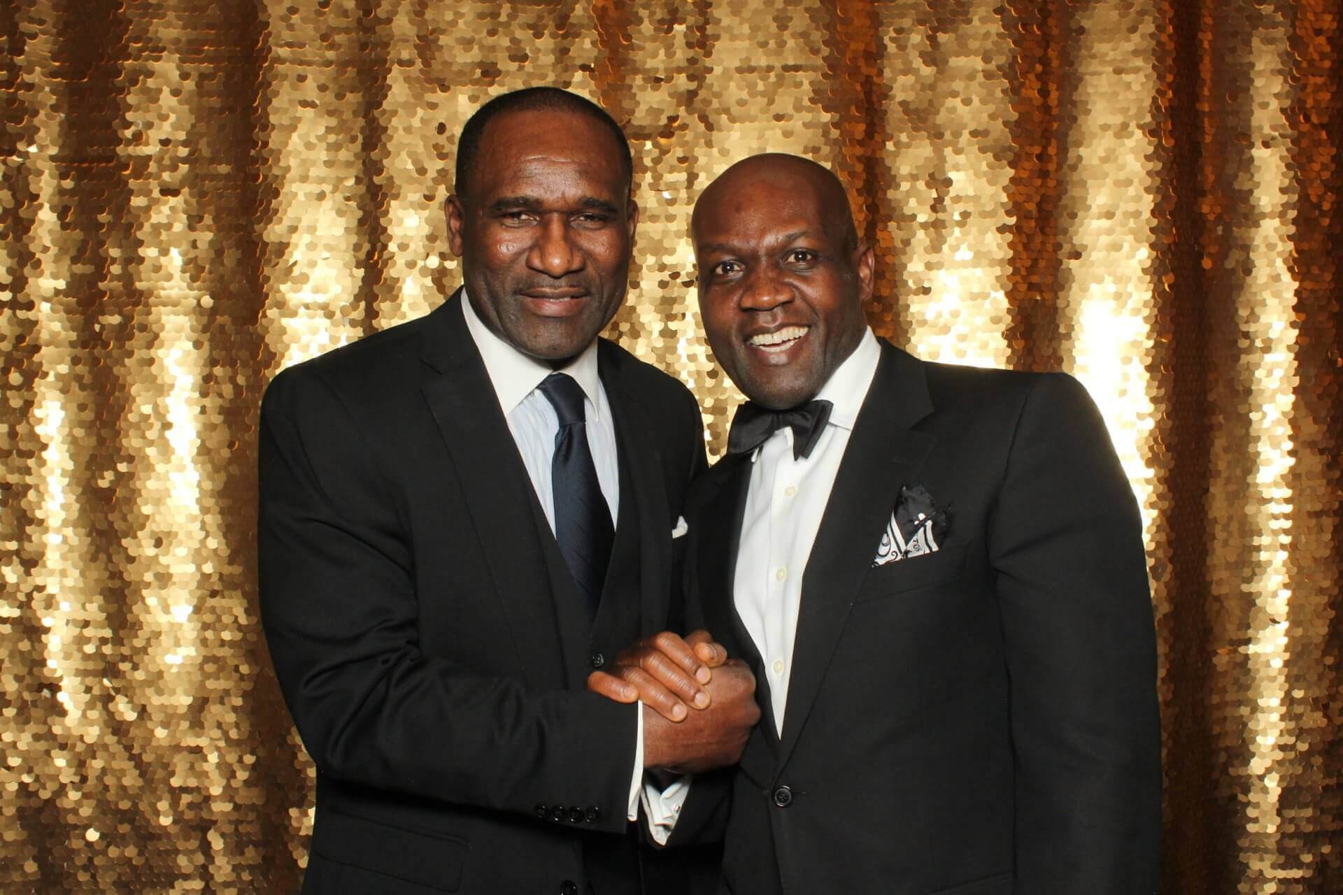 Two men in suits shaking hands in front of a gold sequin backdrop.