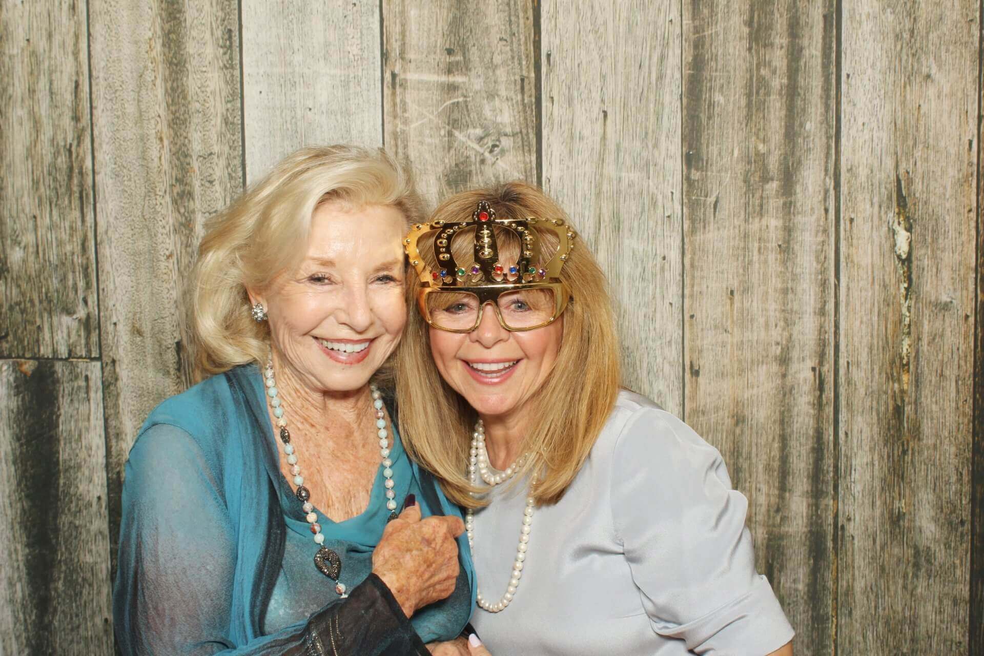 Two women smiling, posing in front of a wooden backdrop. One wears a crown prop, both have necklaces.