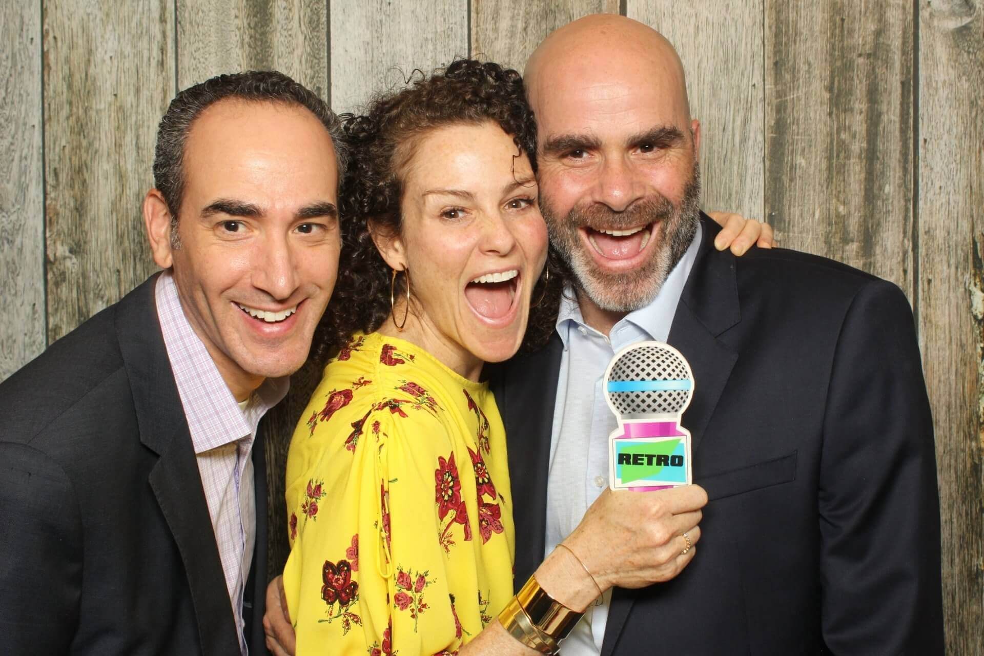 Three people smiling for a photo, one holding a microphone prop in front of a wooden background.