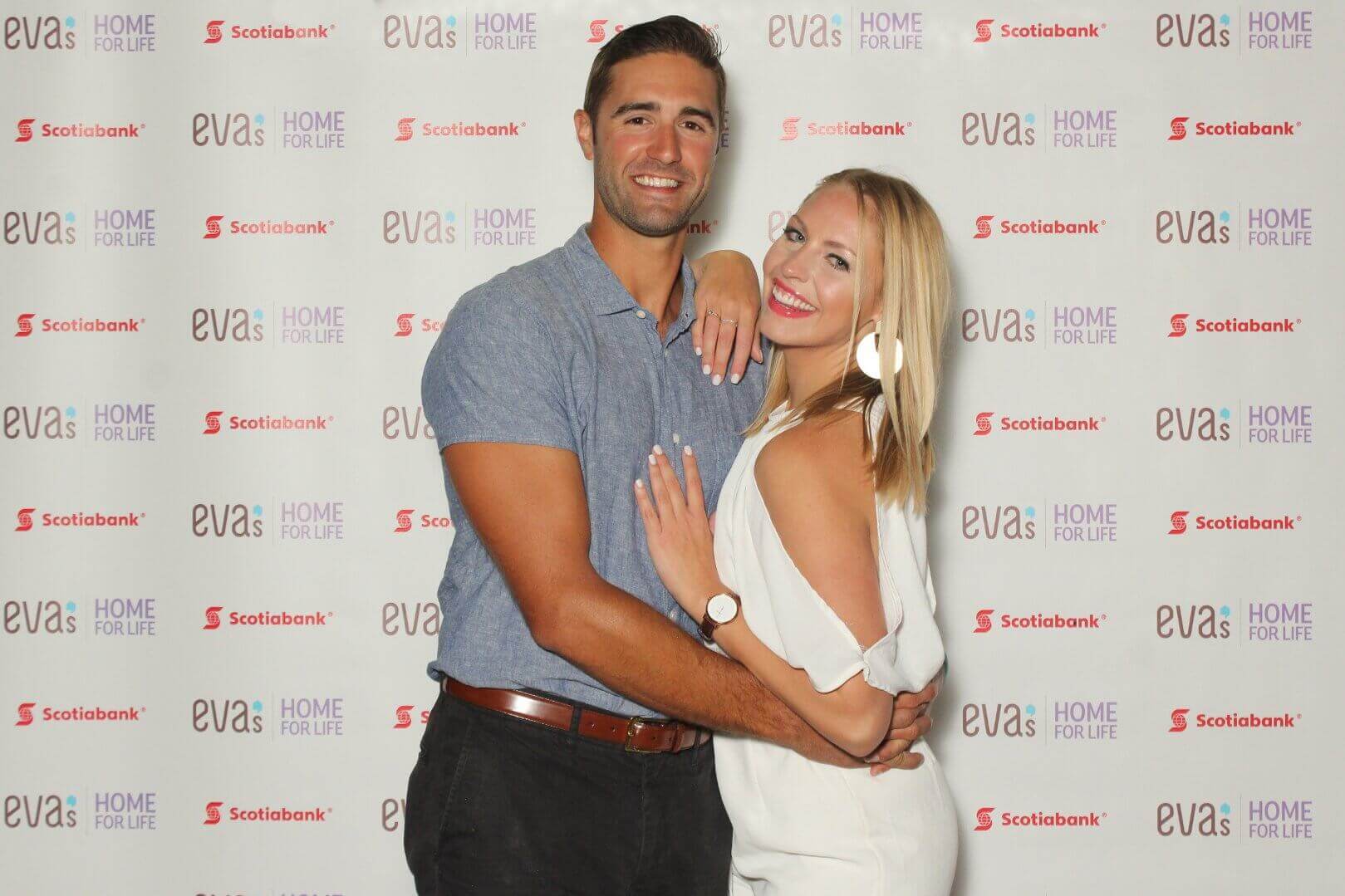 A couple posing in front of a backdrop with logos. The man has his arm around the woman. They are smiling.