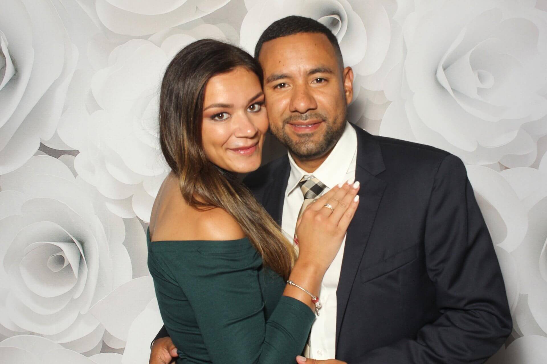 Couple posing closely in front of a white floral backdrop, man in a suit, woman in a green dress, both smiling.