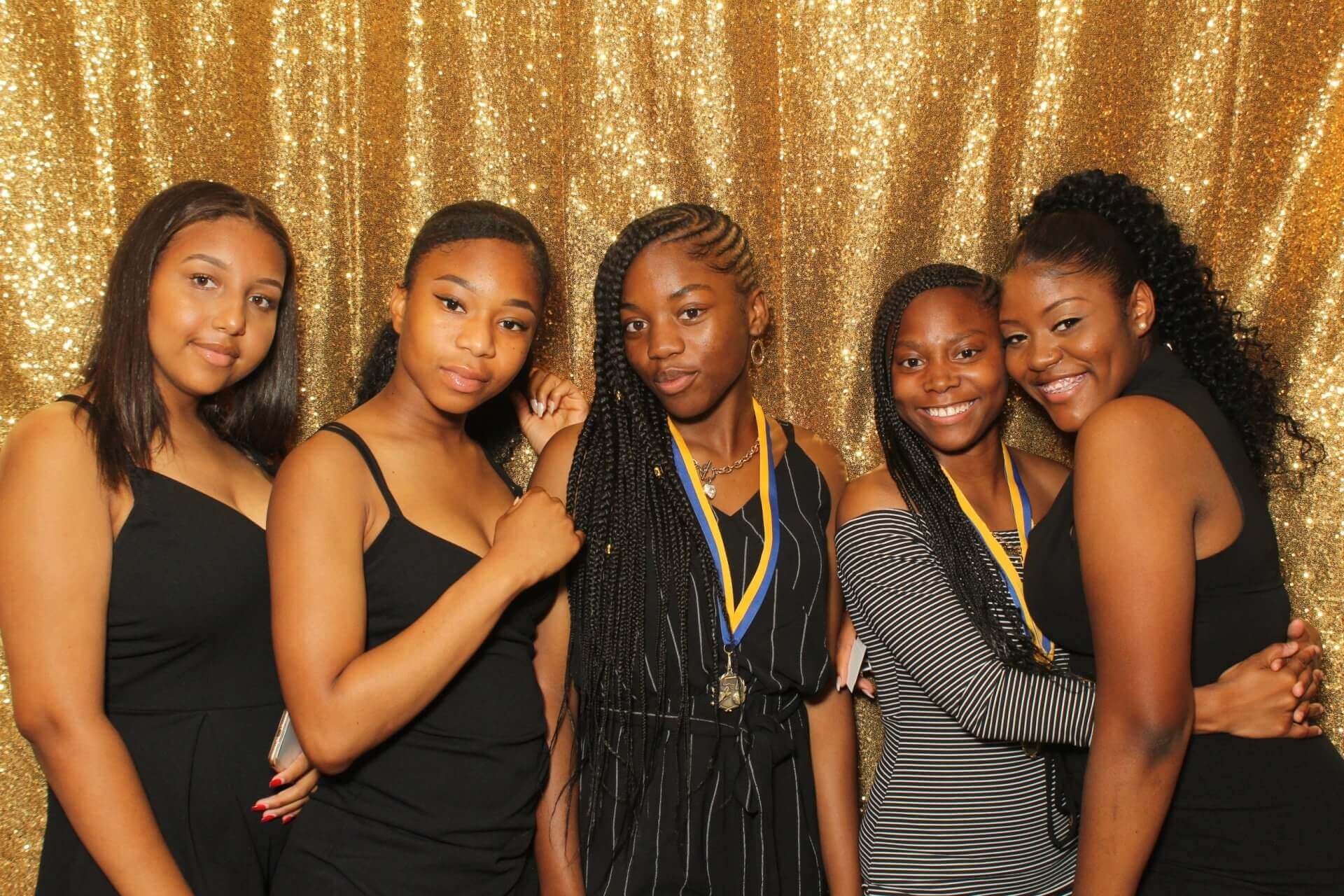 Five people pose for a photo in front of a gold backdrop. They are smiling, wearing dresses, and some have medals.