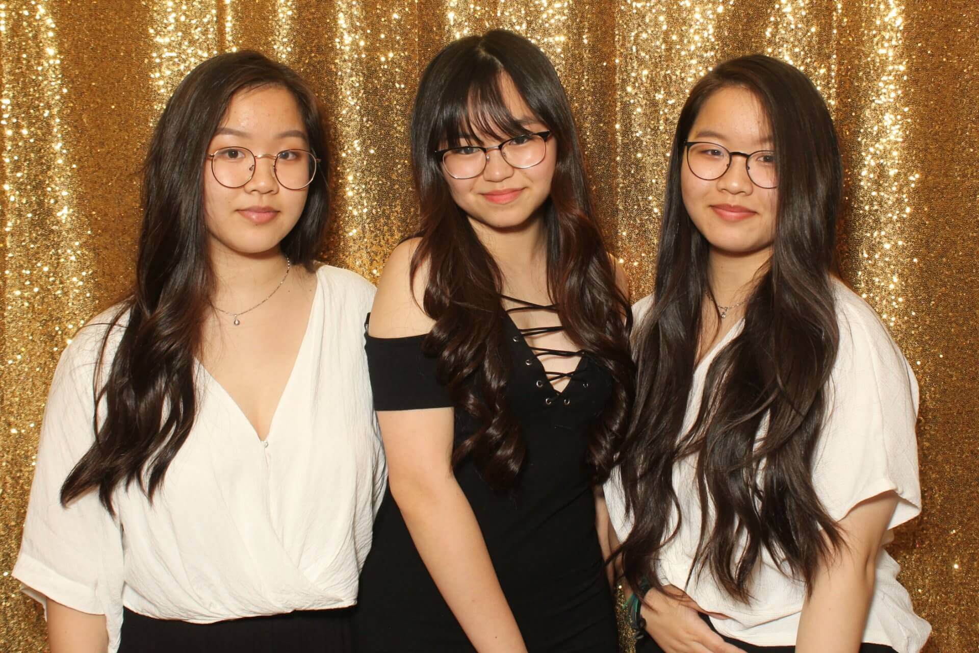 Three young people with glasses pose in front of a gold sequined backdrop.
