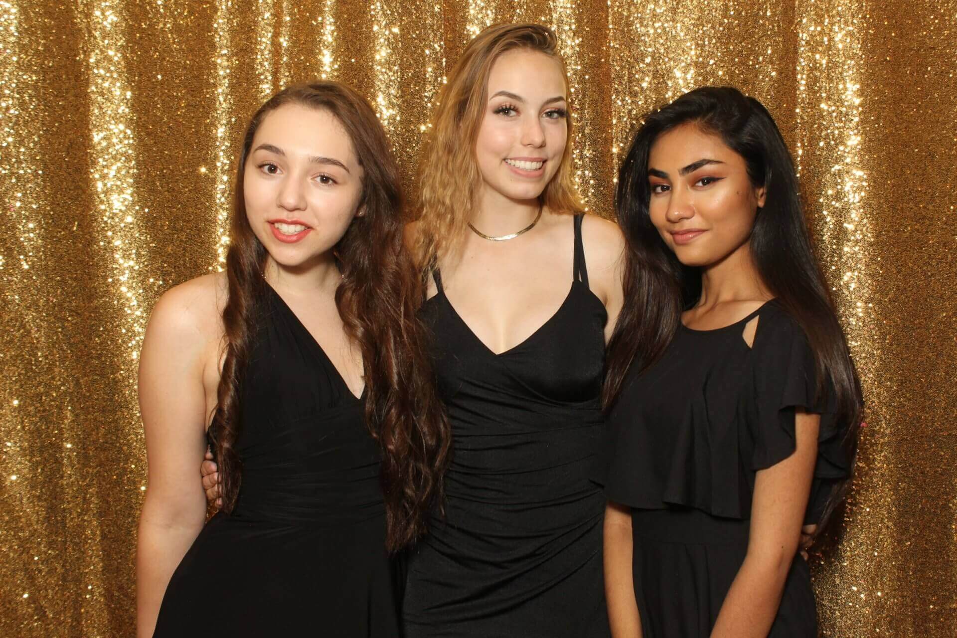 Three young women in black dresses pose in front of a gold sequined backdrop.