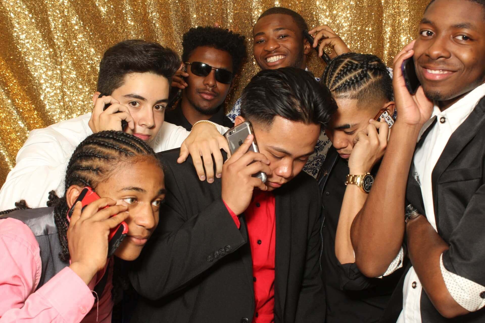 Group of people at an event using cell phones. Gold sequin backdrop. Men in formal wear, some smiling.