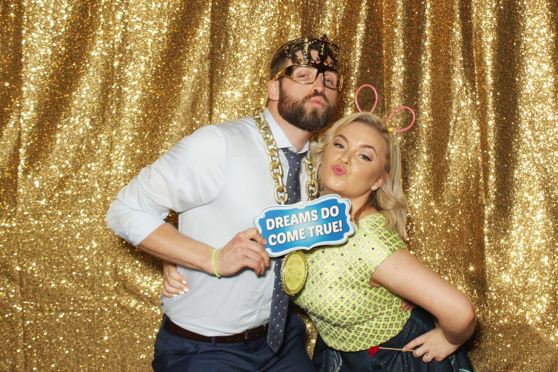 Couple posing at a photo booth; gold backdrop. Man with beard, crown, and sign 
