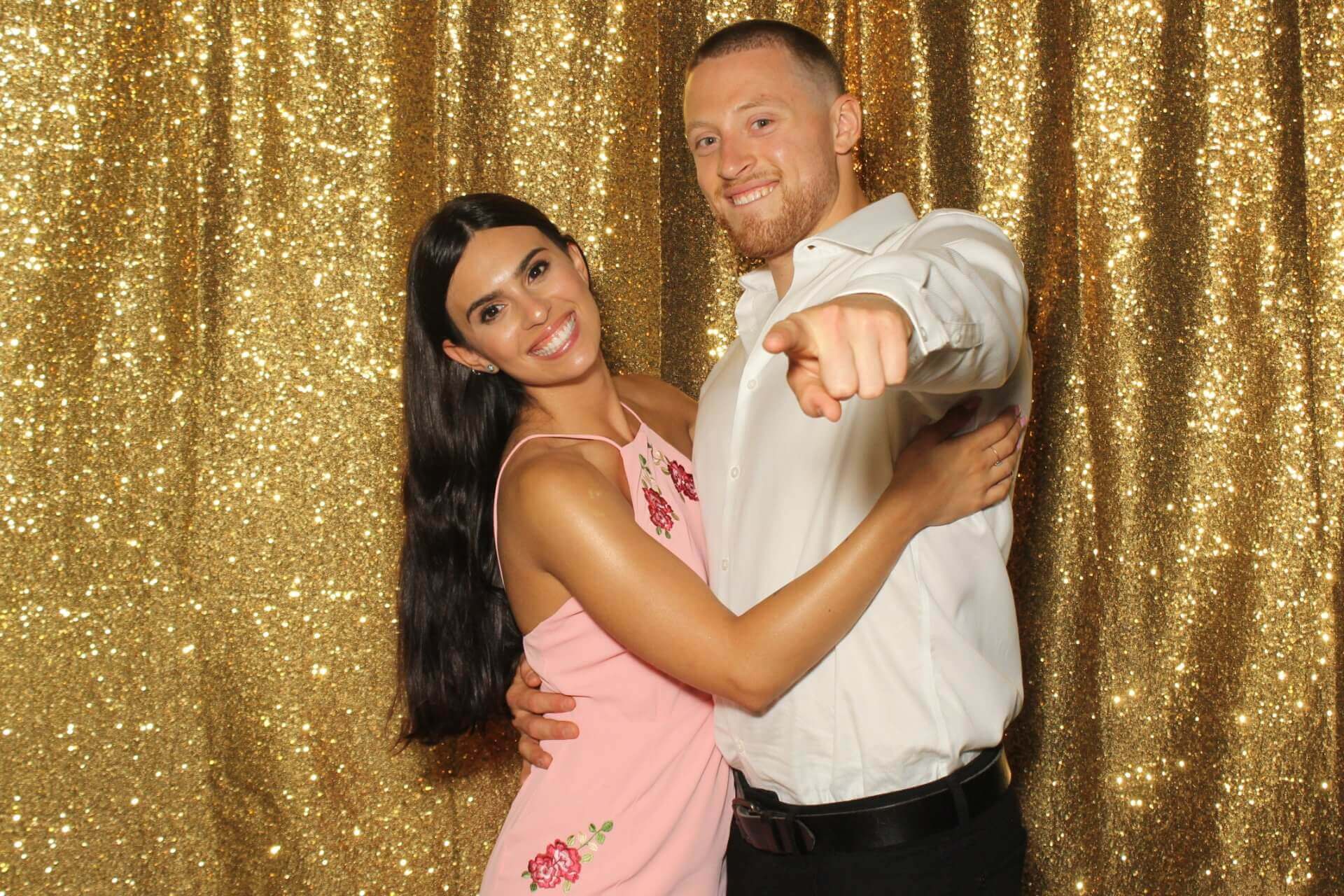 Couple poses for photo booth with gold sequin backdrop; woman in pink dress smiles, man points.
