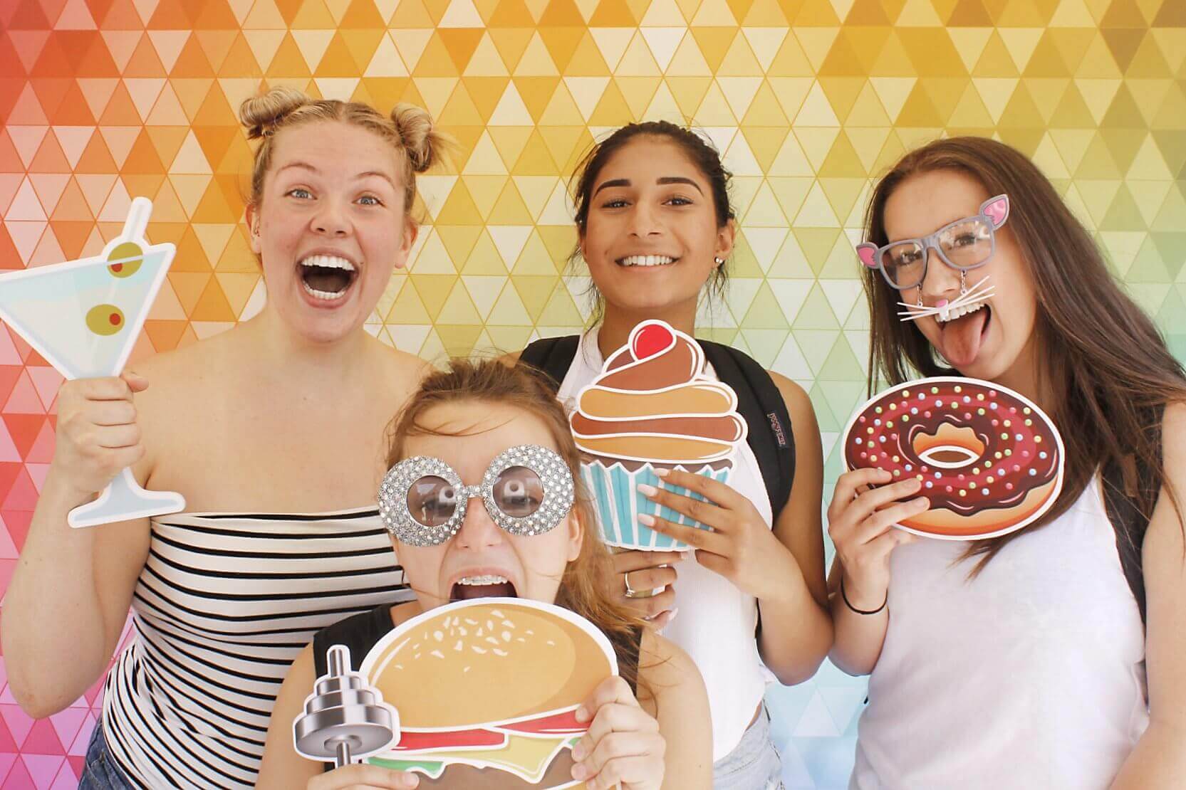 Four people with props, posing in front of a colorful geometric background, all smiling and laughing.