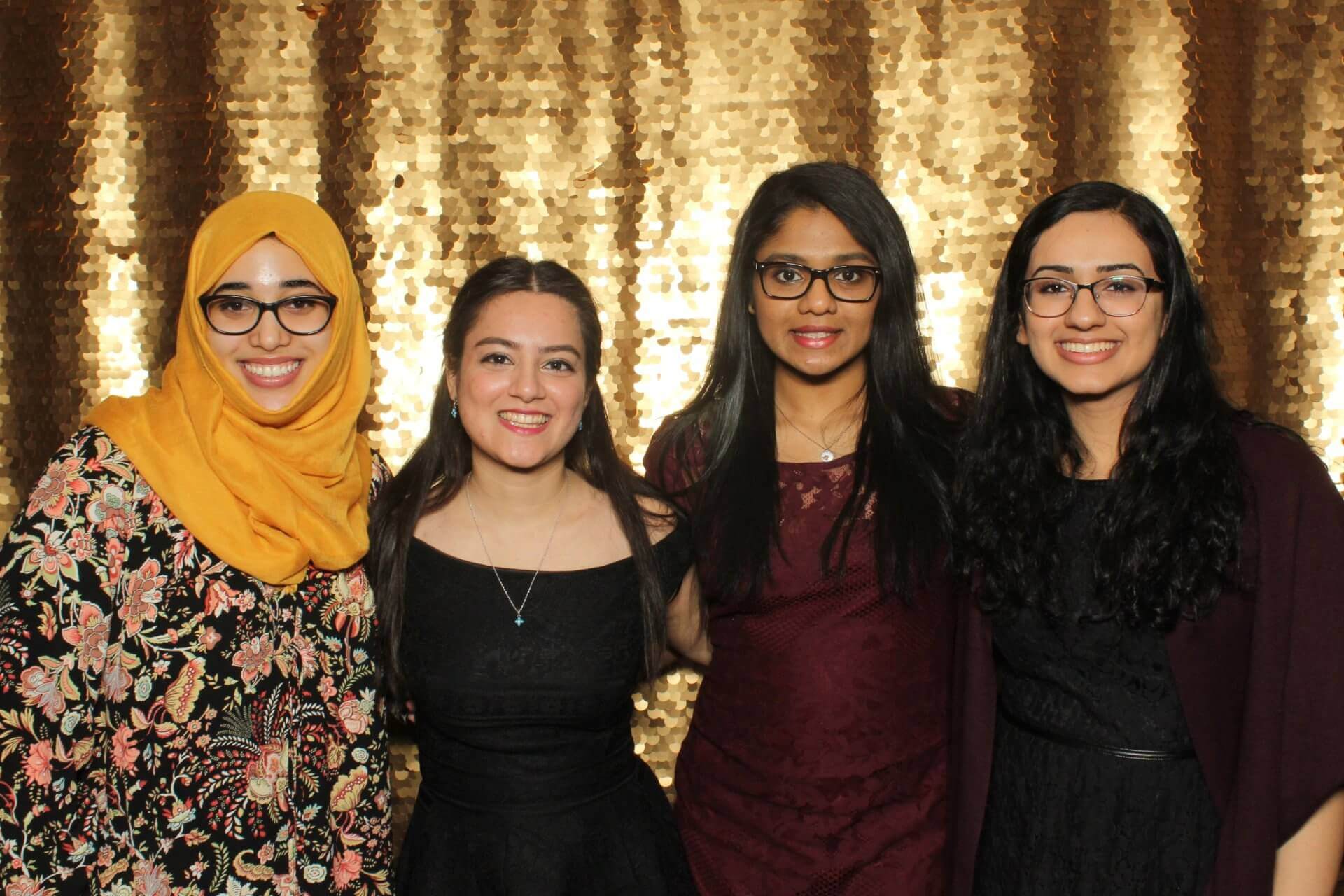 Four women smiling, posing in front of a gold sequined backdrop.
