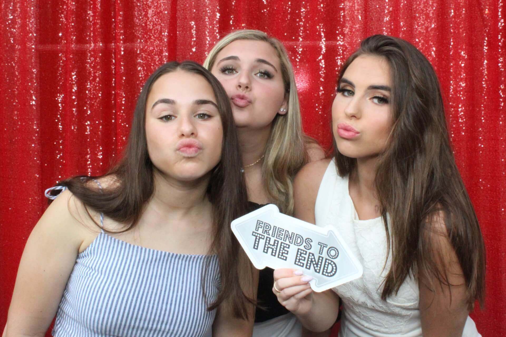 Three young women posing in front of a red sequined backdrop, holding a sign that reads 