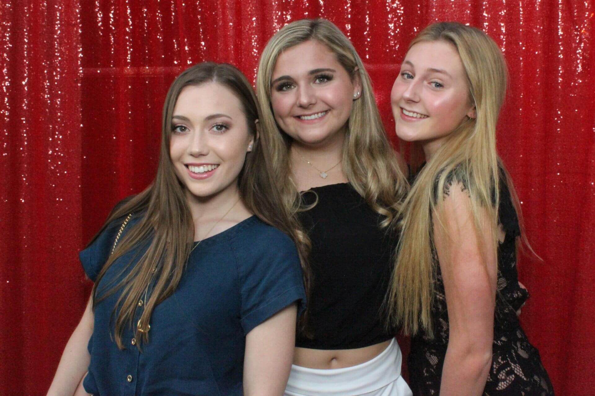 Three young women smiling, posing in front of a red sequined backdrop.
