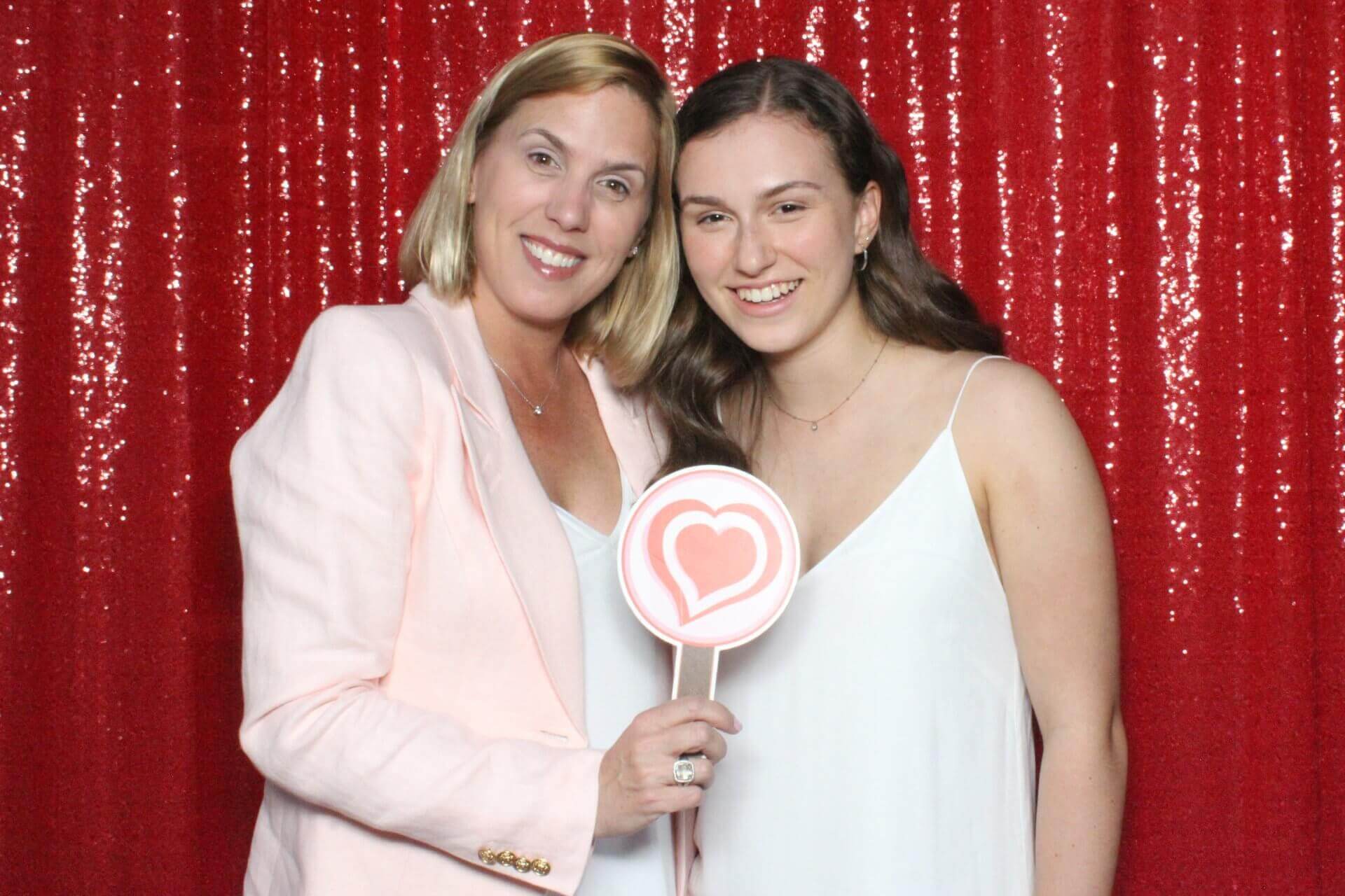 Two women smile, holding a heart-shaped prop, in front of a red sequined backdrop.
