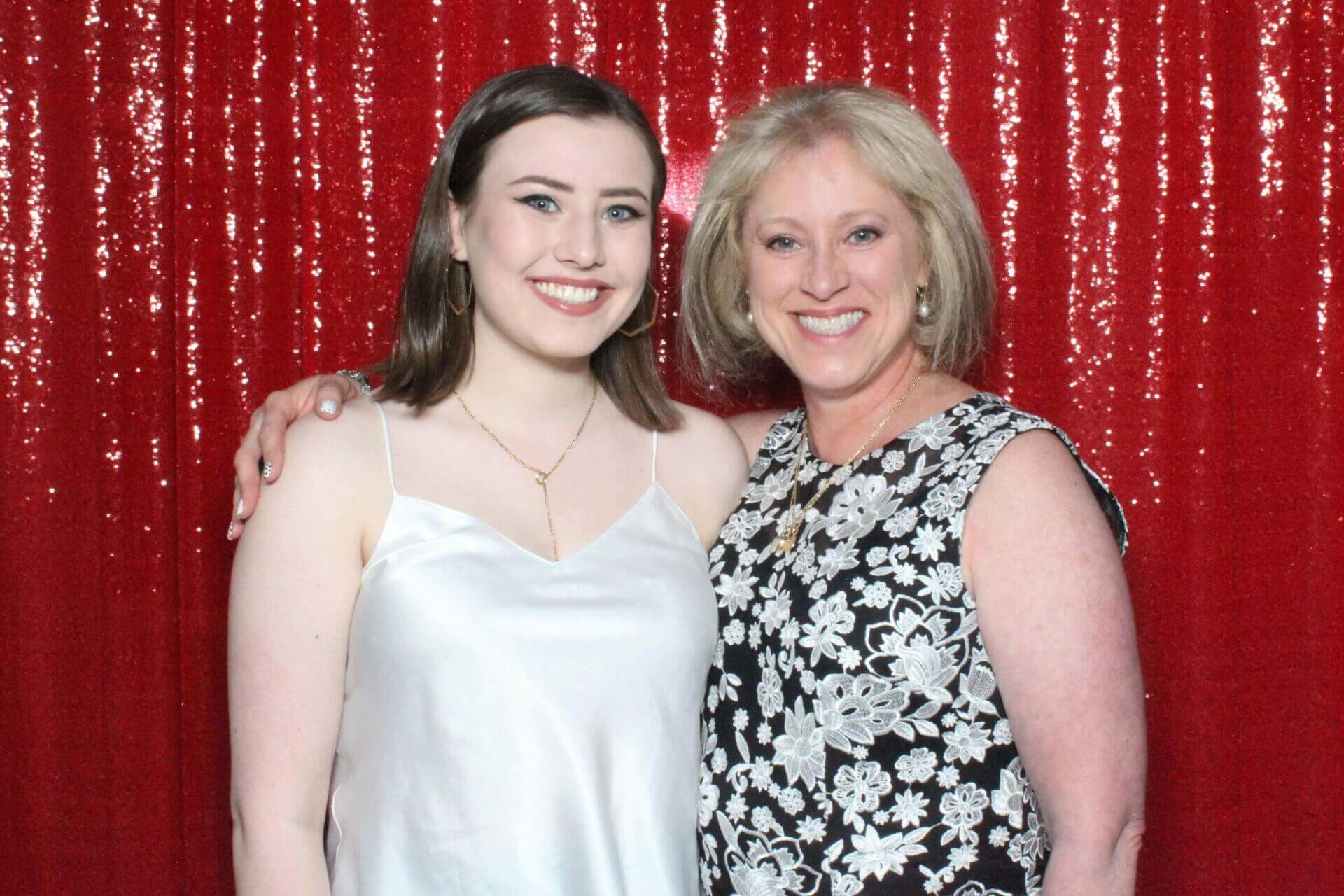 Two smiling people pose against a red sequined backdrop. One person wears a white dress; the other wears a black and white floral top.