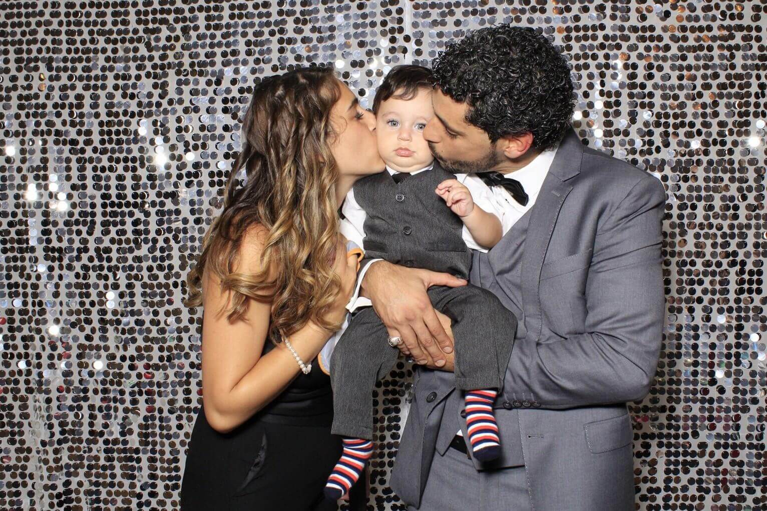 Parents kissing a child in a gray suit and bow tie; shiny silver background.