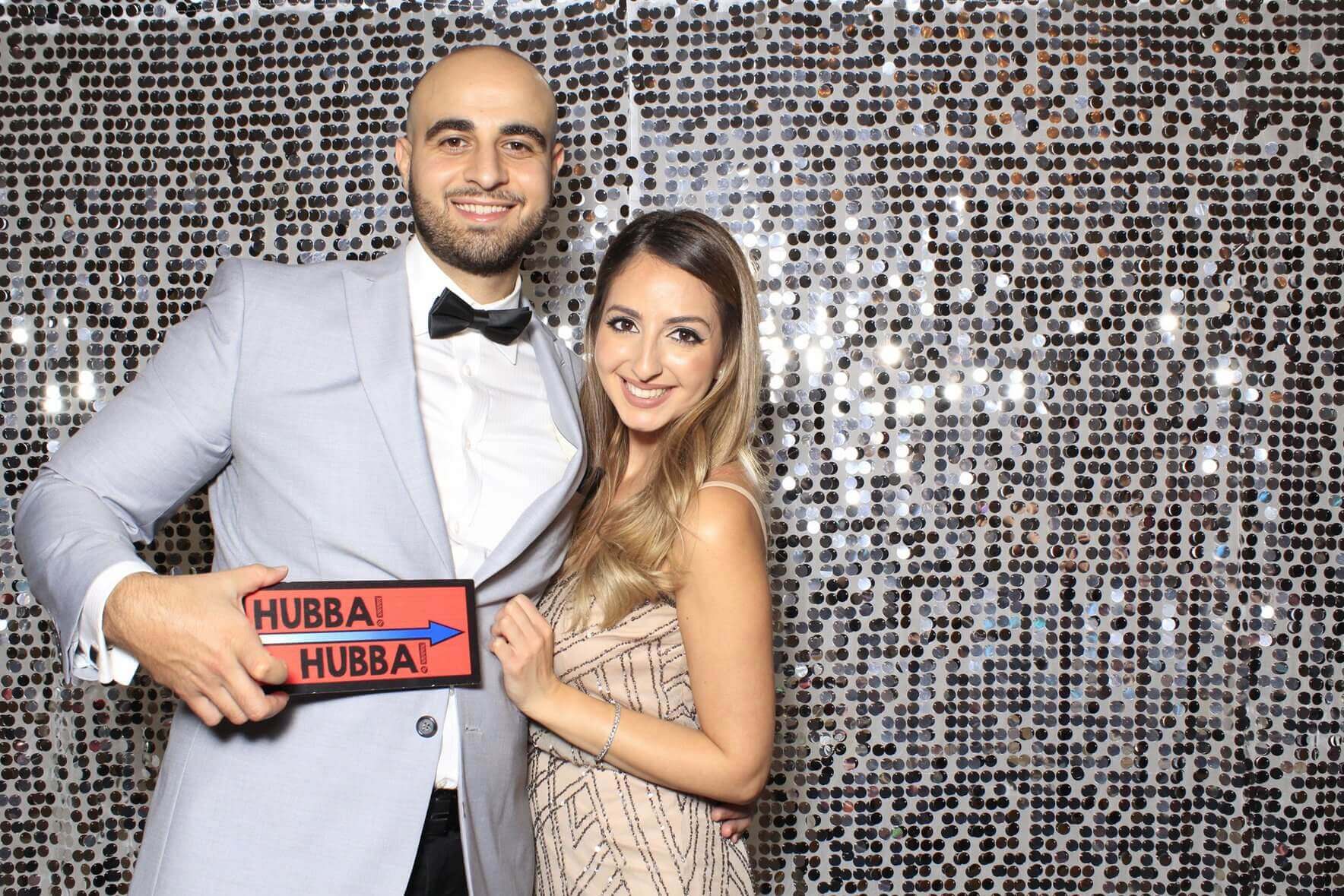 Couple in formal attire smiling, posing in front of a sequined backdrop, holding a 