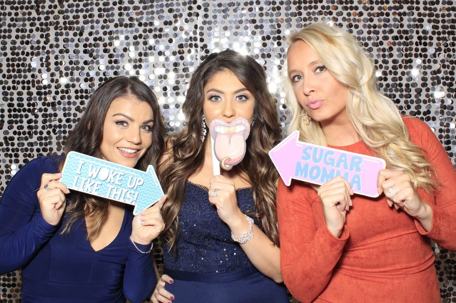 Three women in front of a sequined backdrop pose with novelty signs: 