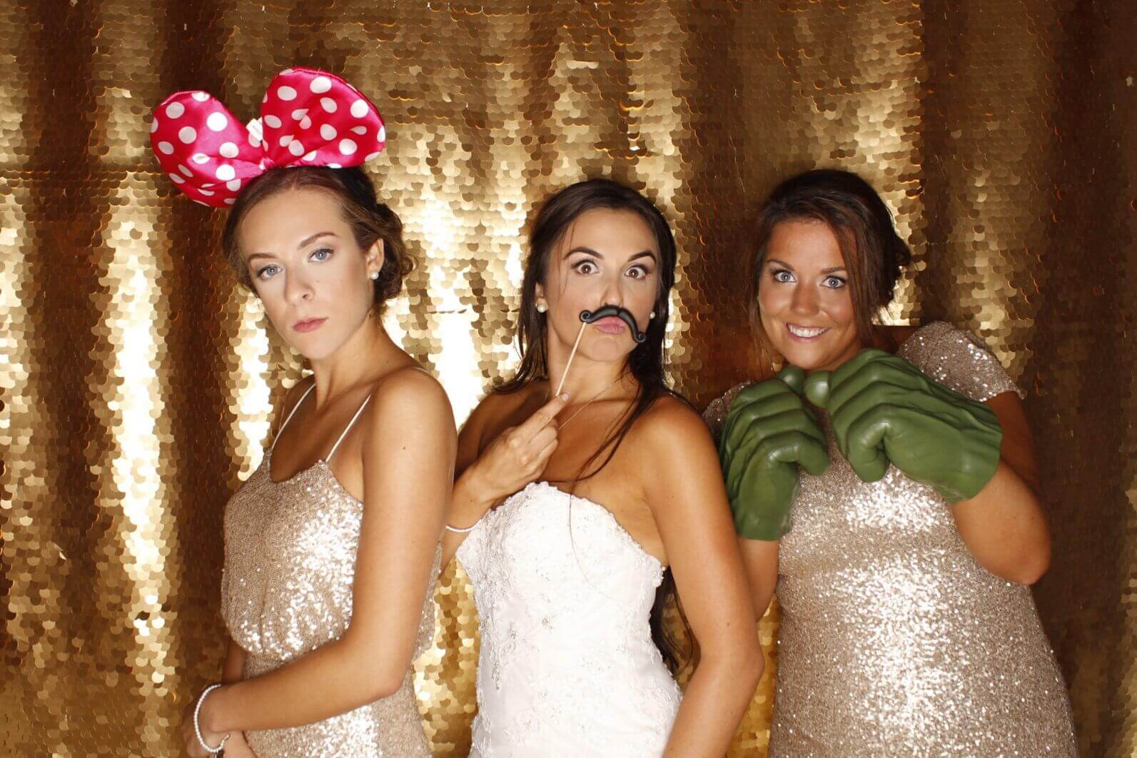 Three women in sequined dresses pose with photo booth props against a gold backdrop.