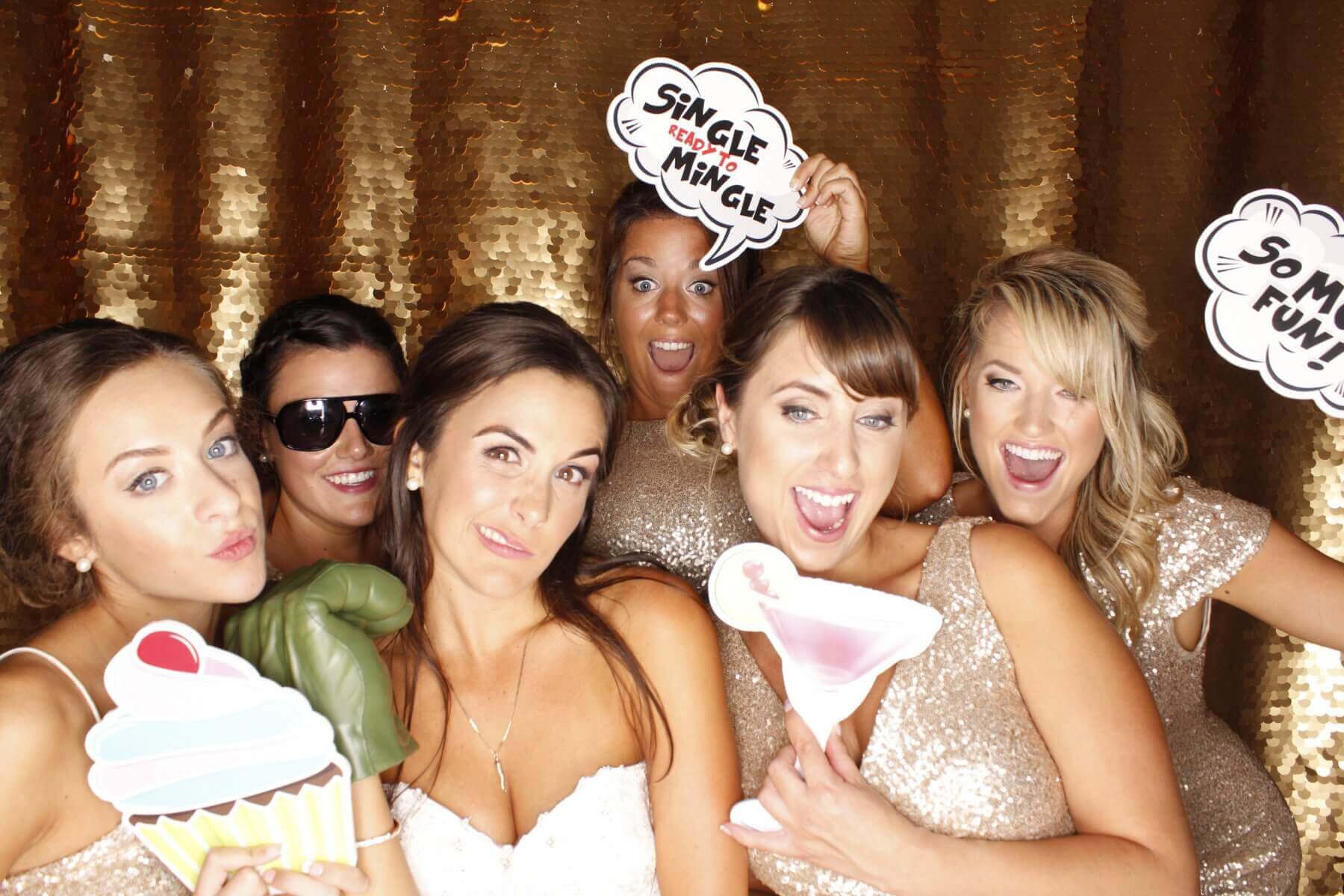 Group of women in gold dresses in a photo booth, holding props, smiling and posing against a gold sequined backdrop.