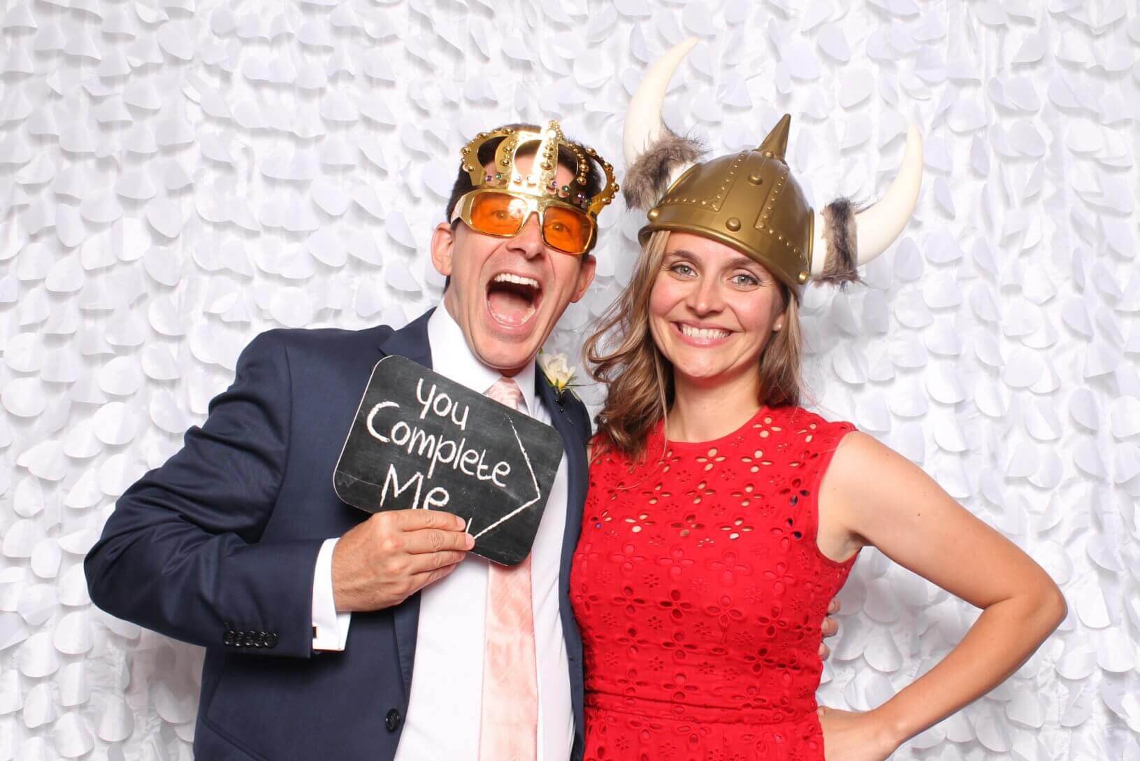 Man in a suit and woman in a red dress posing with fun hats and a chalkboard that says, 
