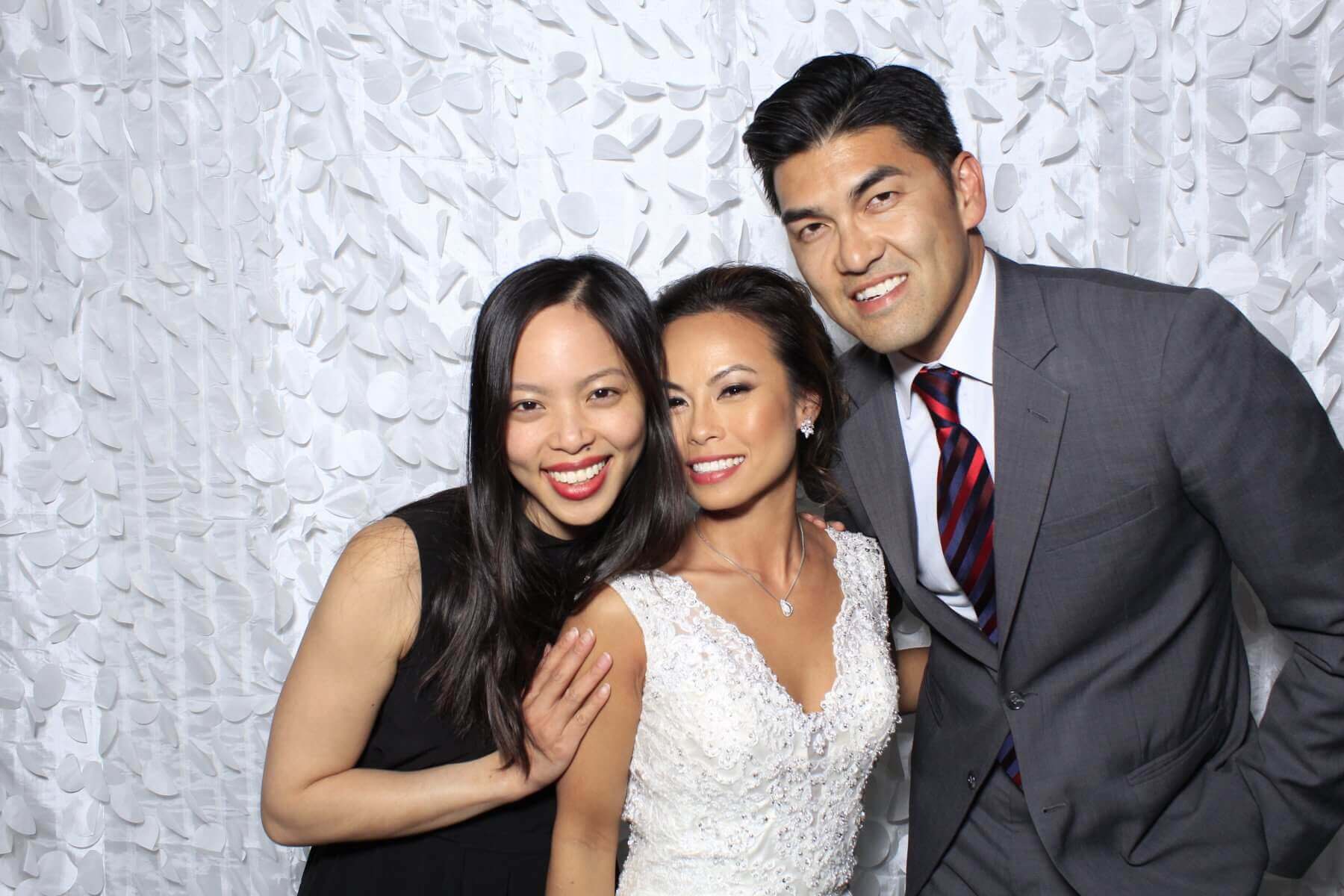 Three people smiling for a photo; one in a wedding dress, one in a suit, and one in a black dress against a white background.