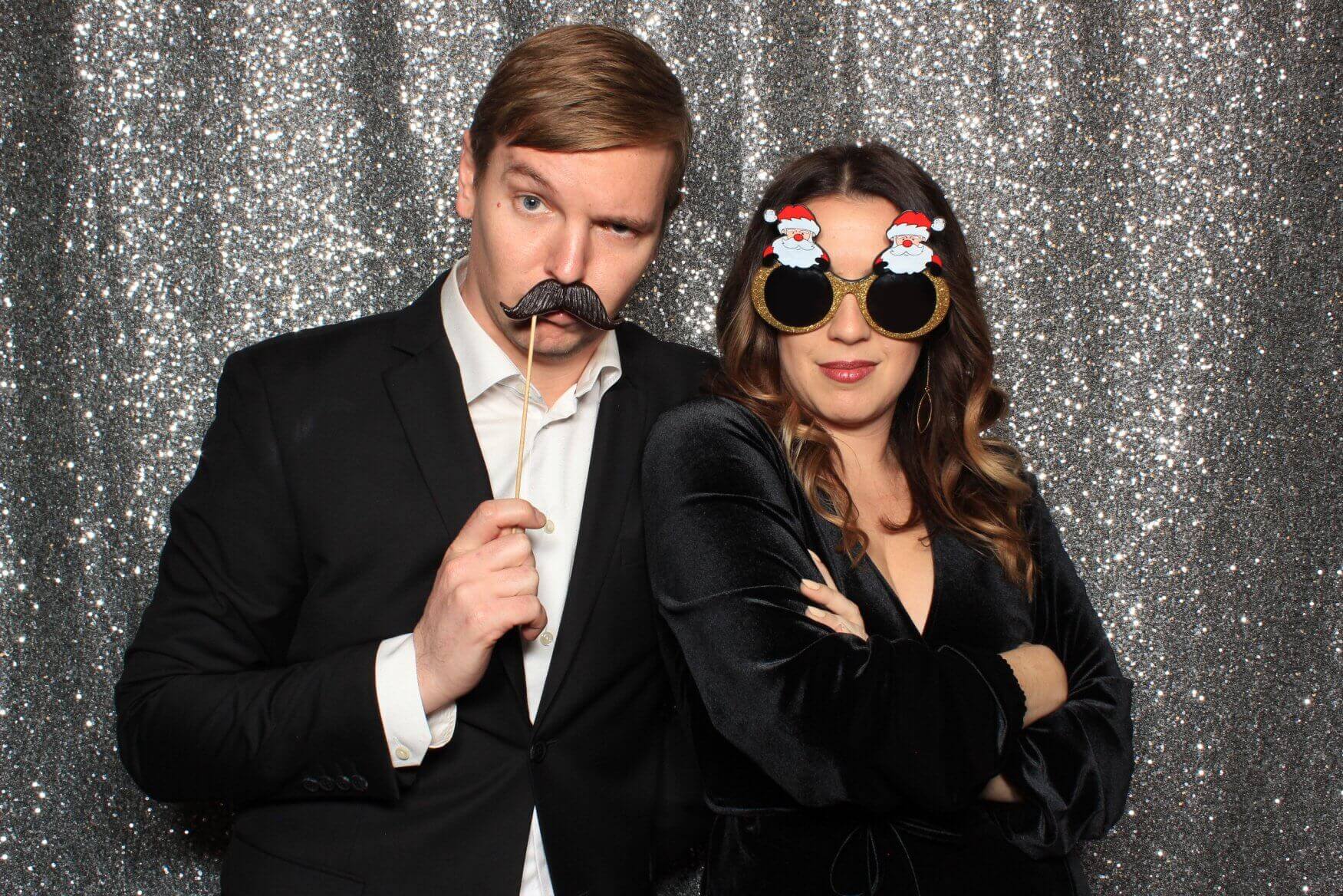 Man holding mustache prop, woman wearing Santa glasses, posing in front of a sparkly backdrop.