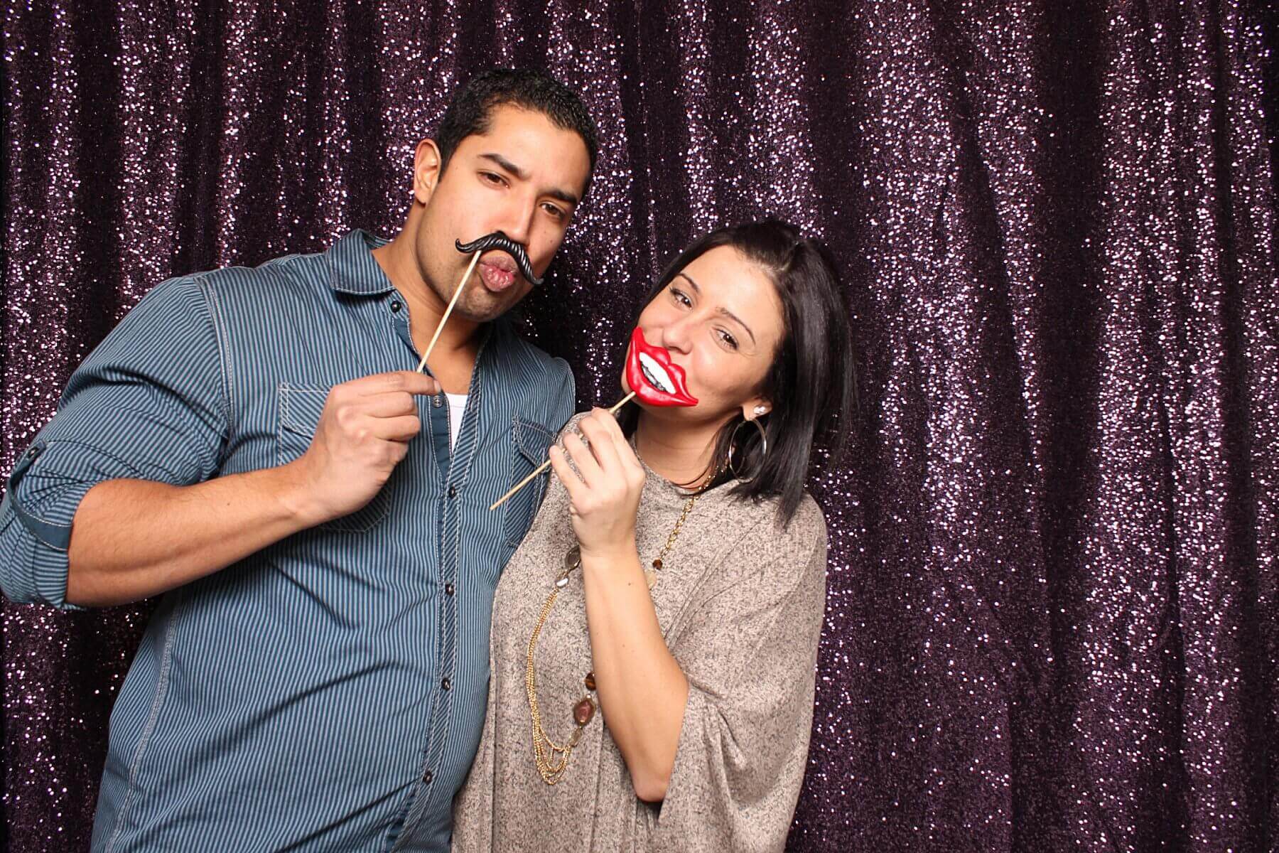 Couple posing with photo booth props in front of a sparkly curtain. Man holds mustache, woman holds lips.