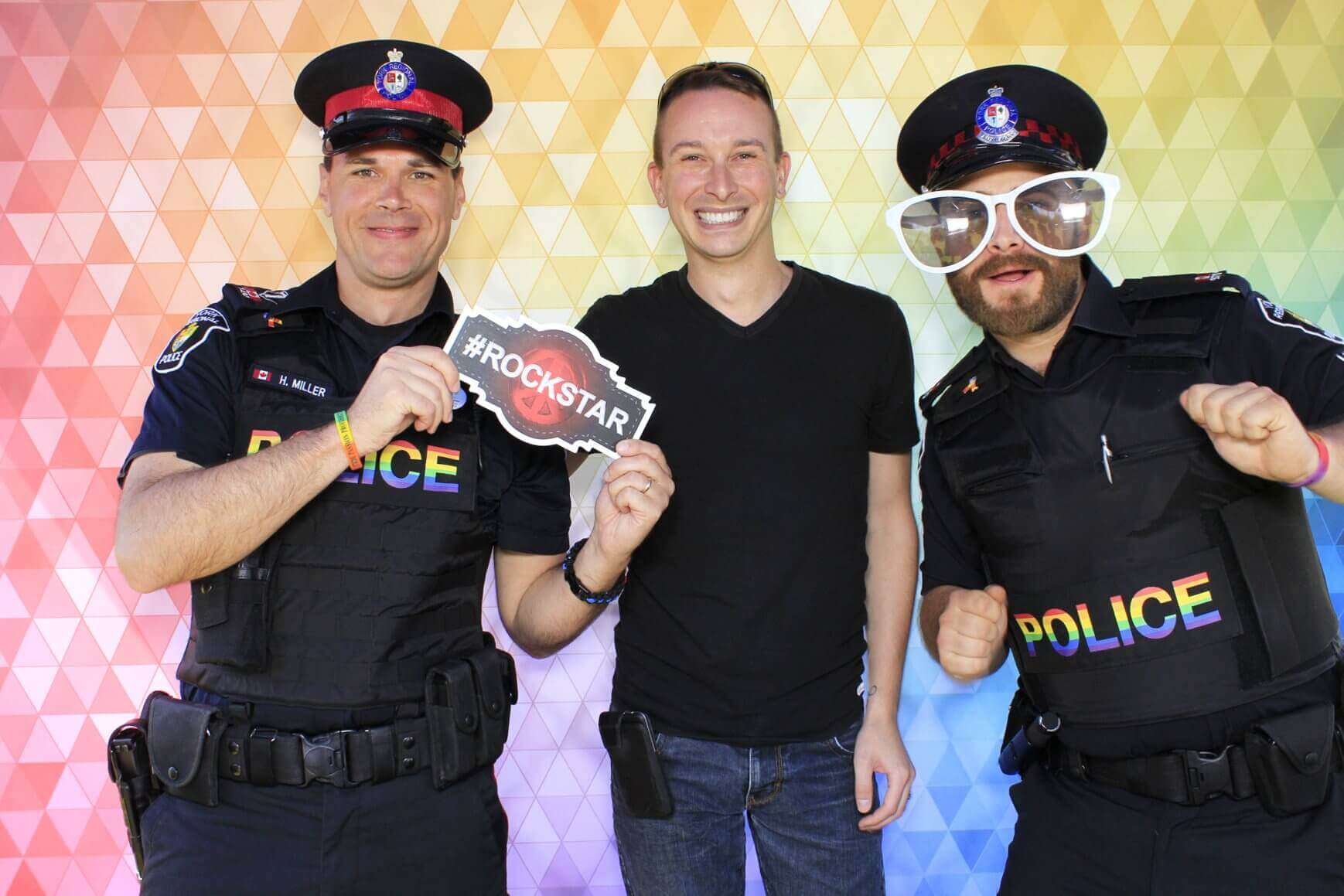 Two police officers and a civilian pose in front of a rainbow backdrop. One officer wears novelty glasses, all smiling.