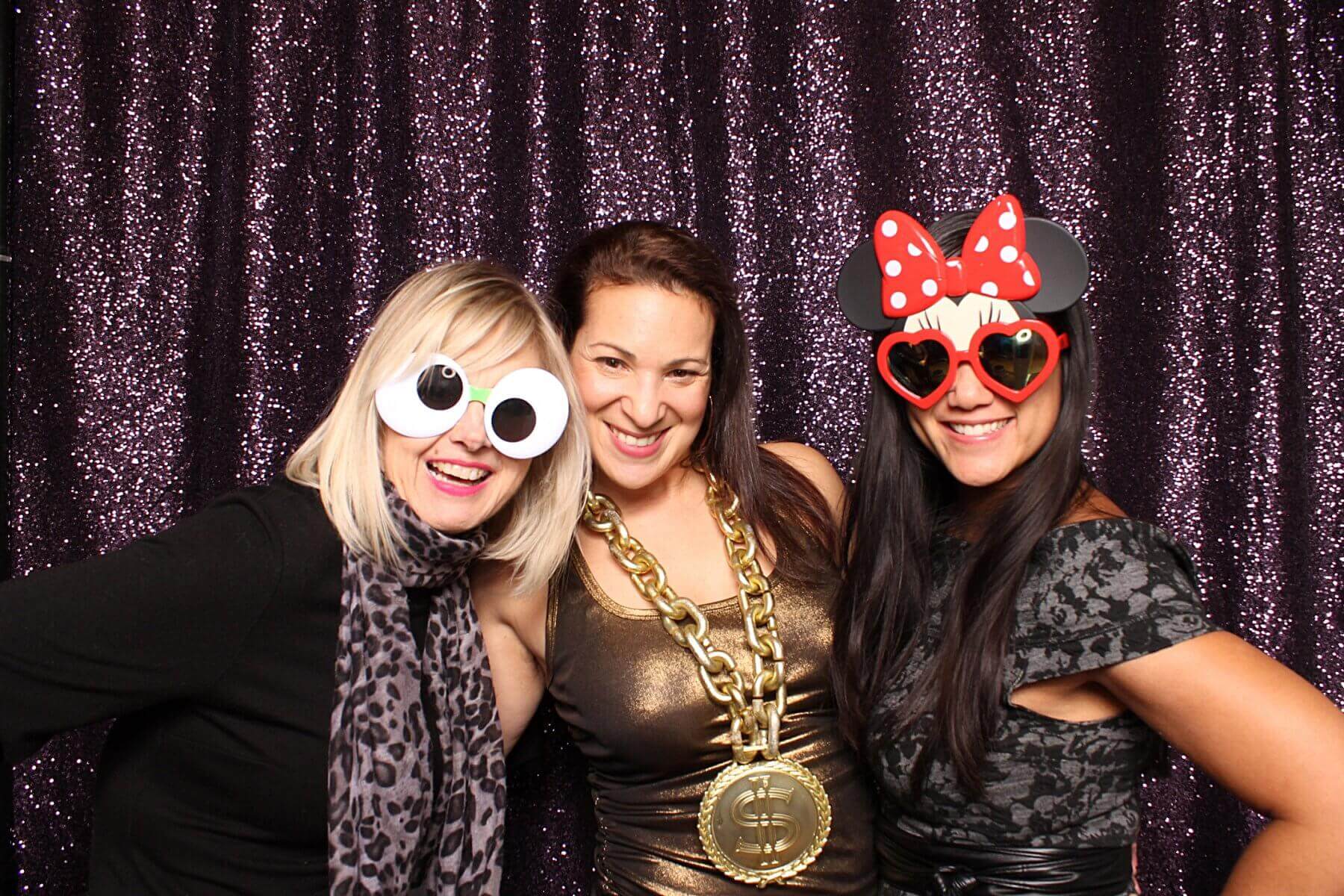 Three smiling women pose at a photo booth with props. One wears googly-eye glasses, another a gold chain, and the third a Minnie Mouse headband.