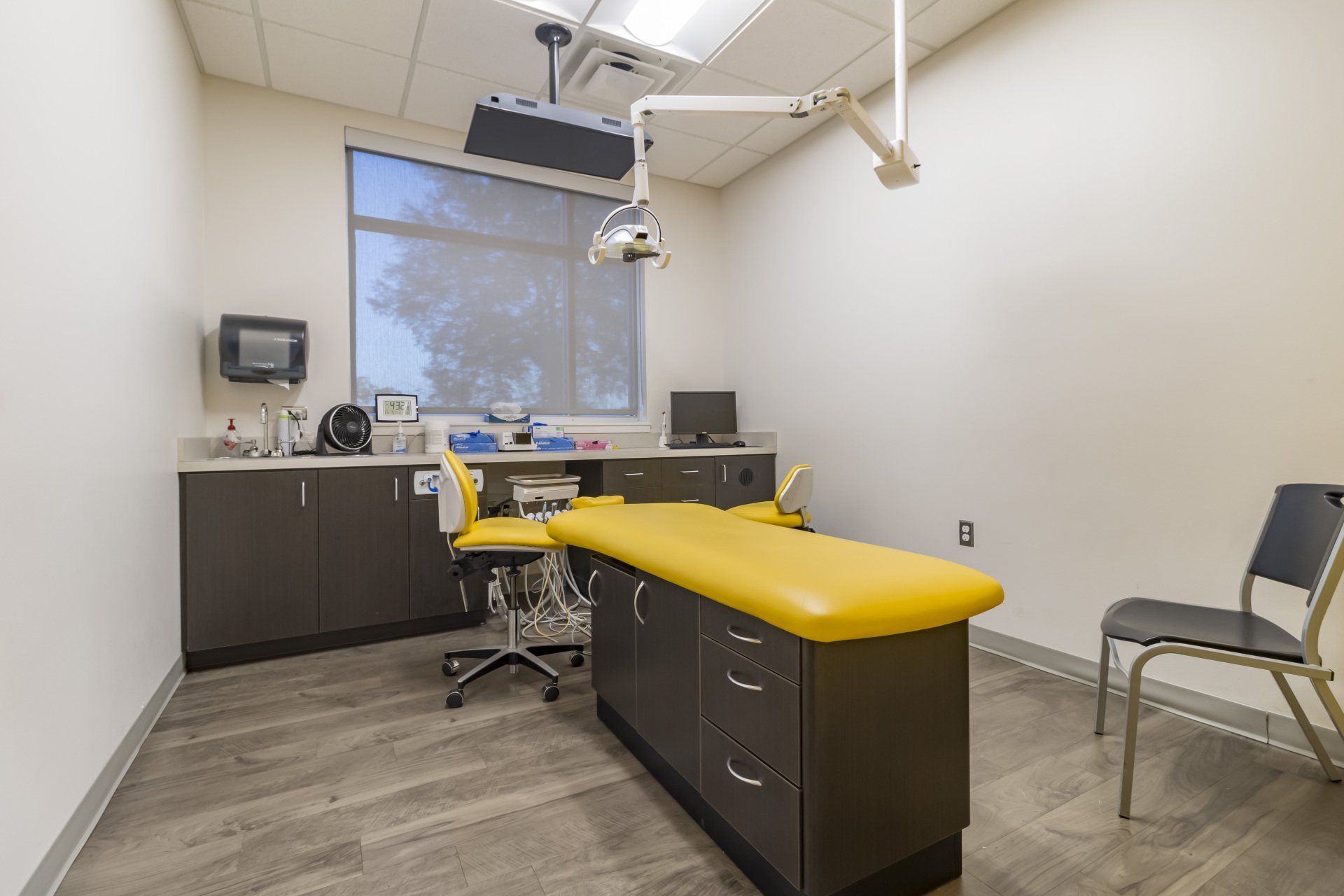 Examination room with yellow examination table, gray cabinets, surgical light, and a window.