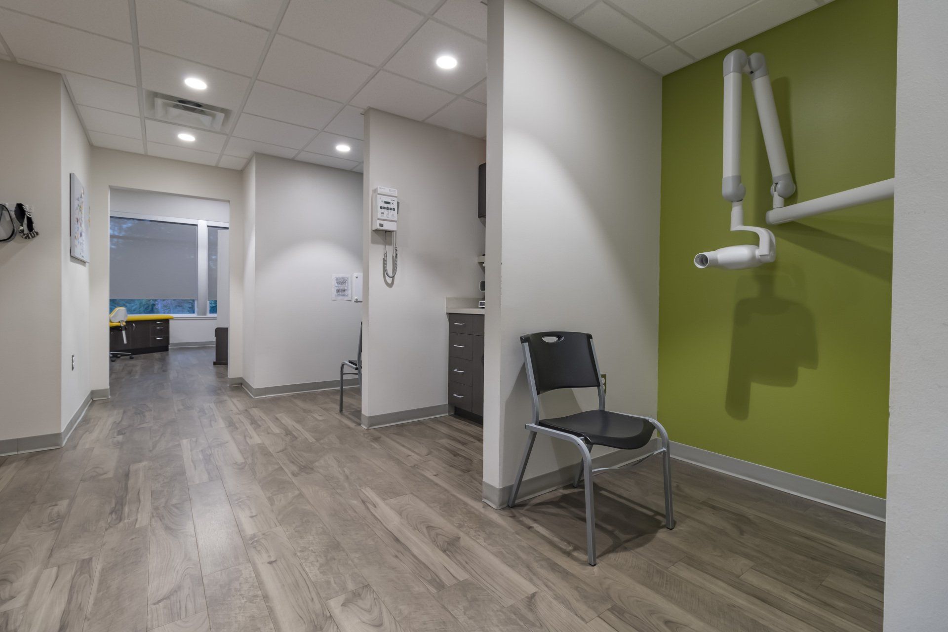 Dental office hallway with light gray wood-look flooring, light walls, and a green accent wall with an X-ray machine.