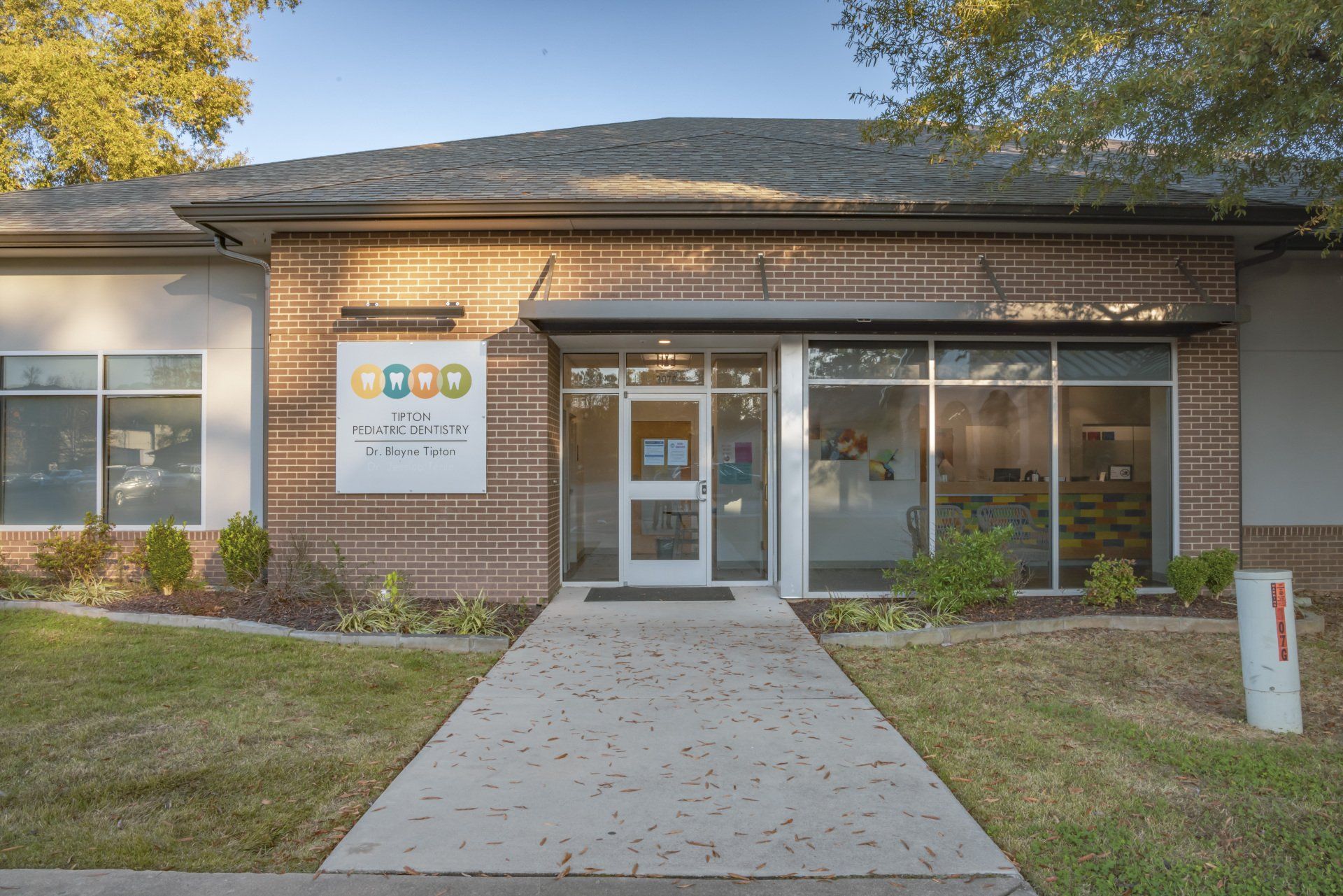 Exterior of a brick building with a walkway leading to a glass door entrance. Green lawn surrounds.