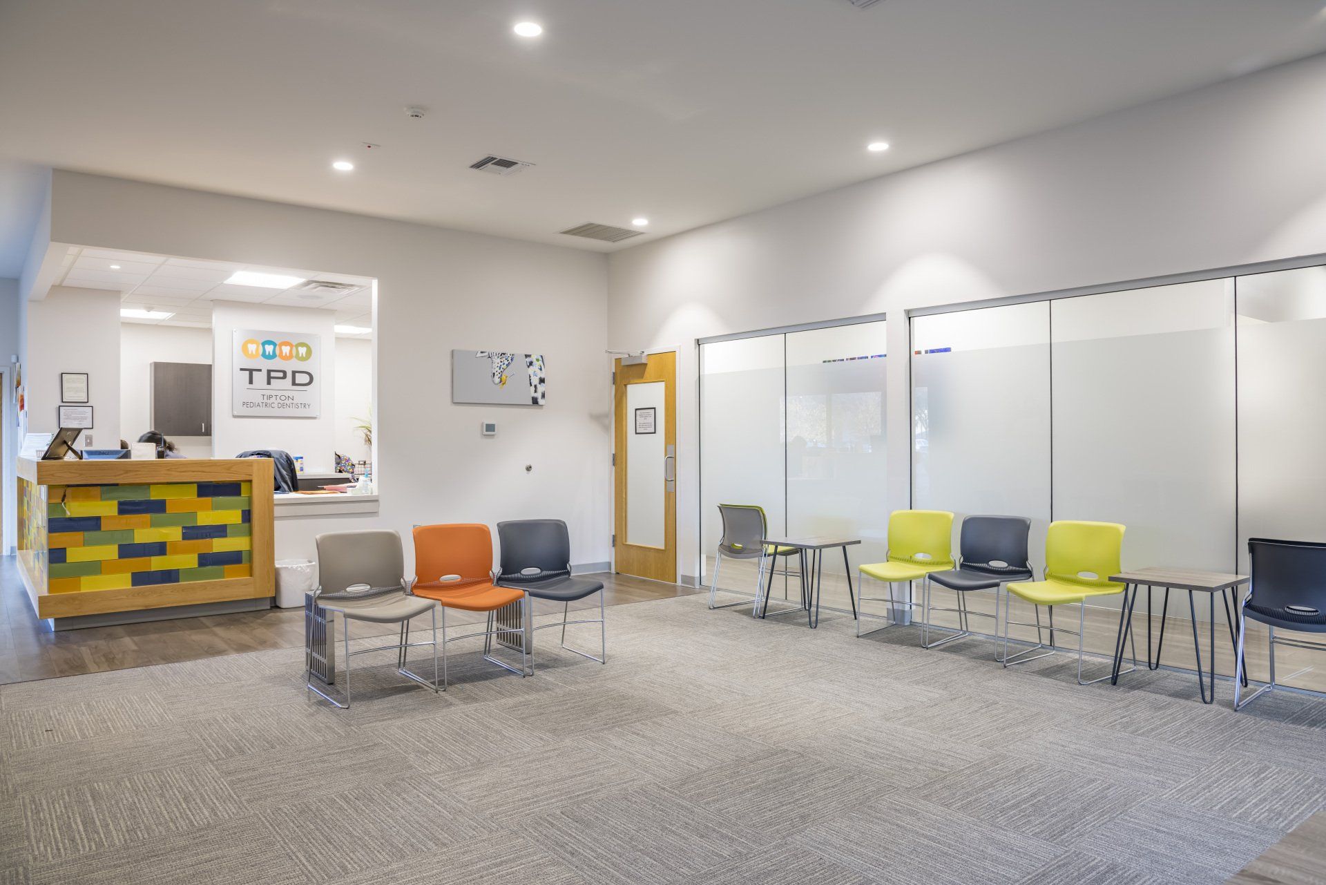 Waiting room with colorful chairs, rug, and frosted glass walls. Reception desk visible.