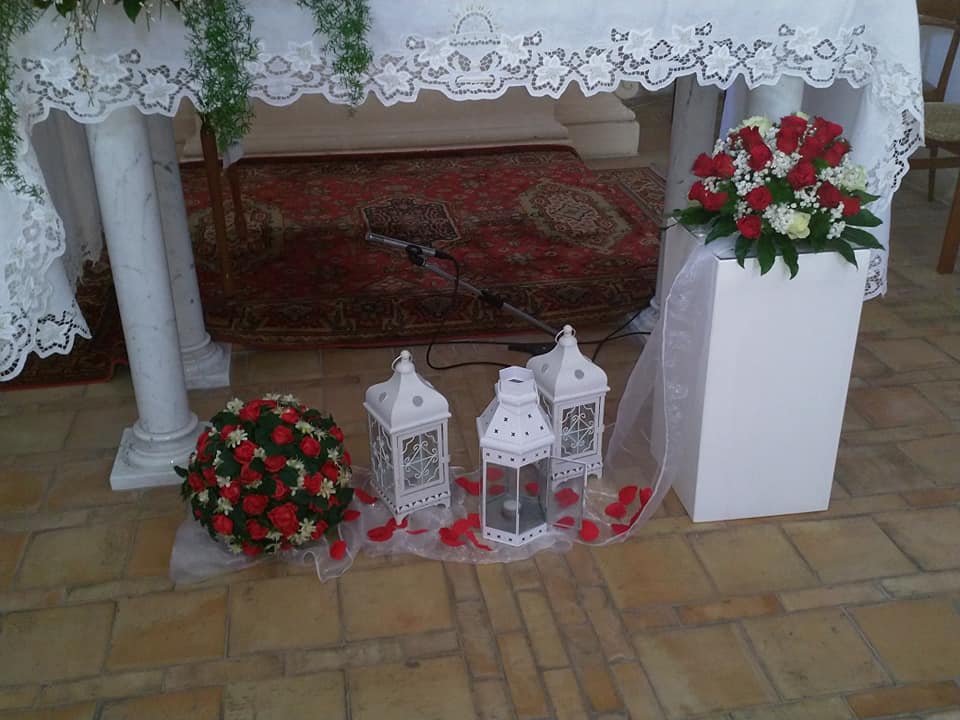 Altare con fiori, lanterne e piedistallo bianco. Rose rosse e pizzo bianco decorano l'ambiente.