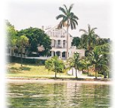 A large white house is sitting on the shore of a lake surrounded by palm trees.