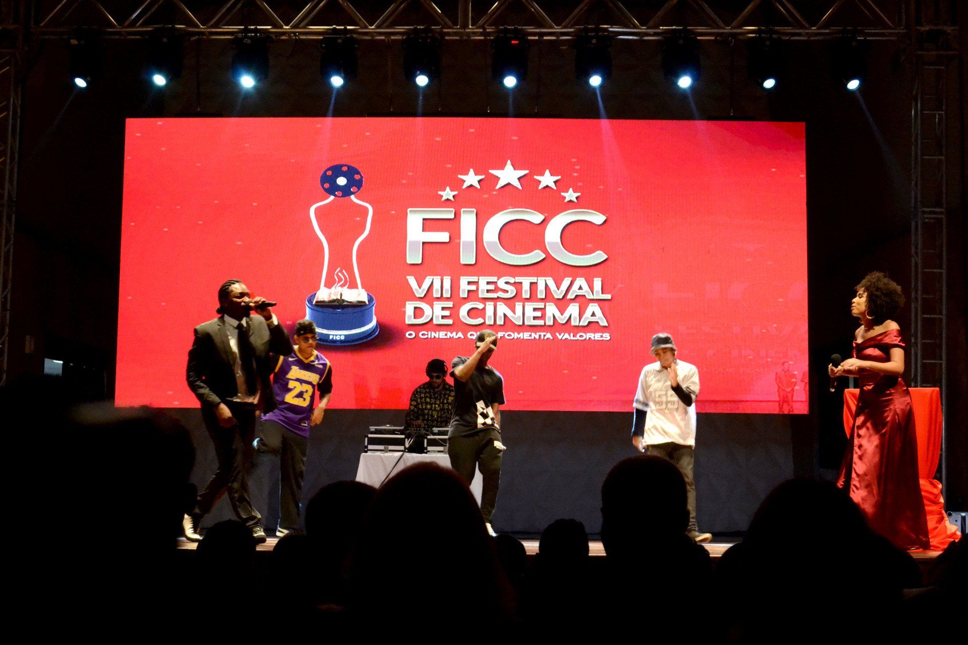 Um grupo de pessoas está em pé em um palco em frente a uma tela grande que diz ficc festival de cinema