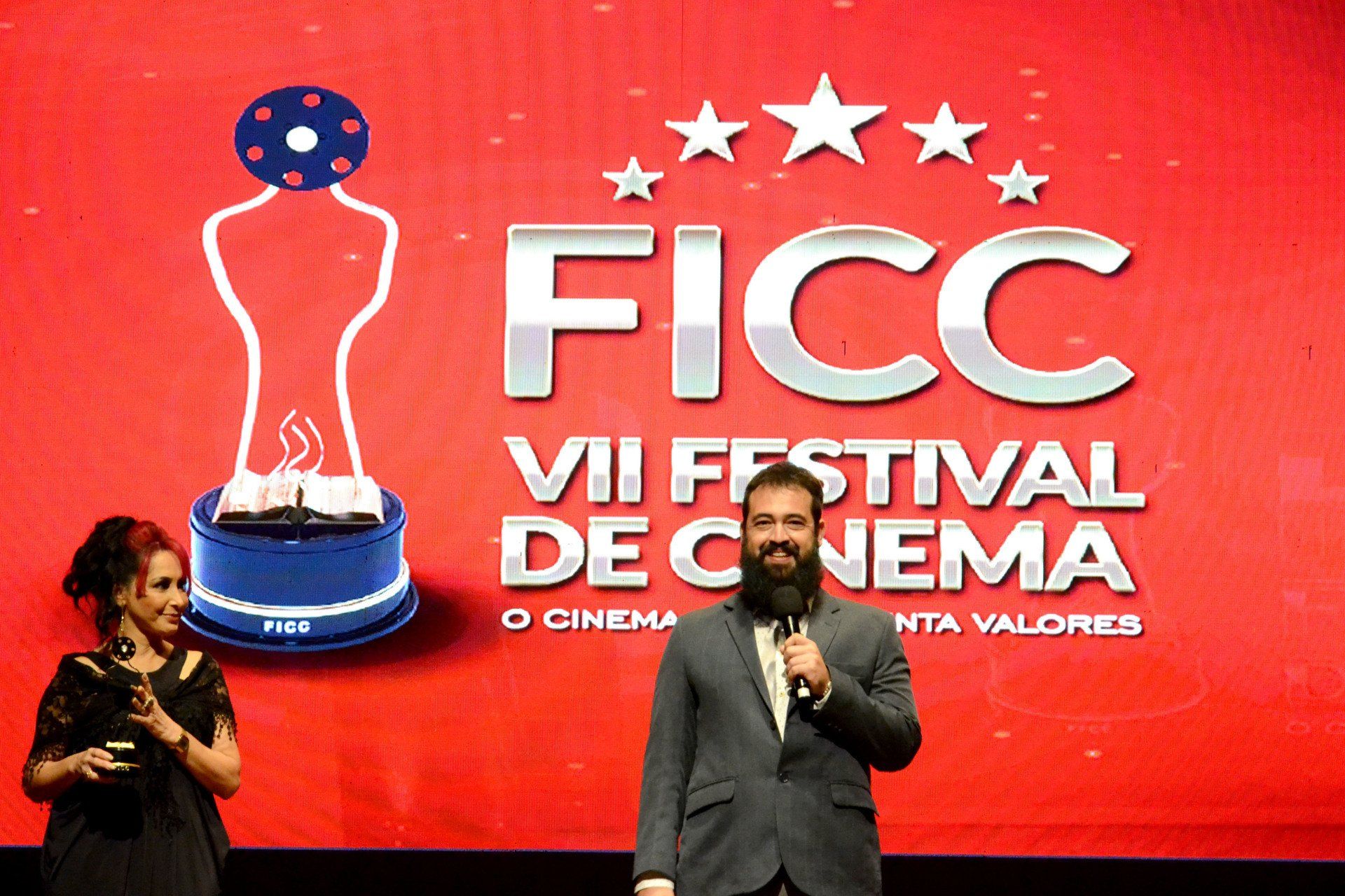 Um homem e uma mulher estão em pé em frente a uma tela que diz ficc vii festival de cinema