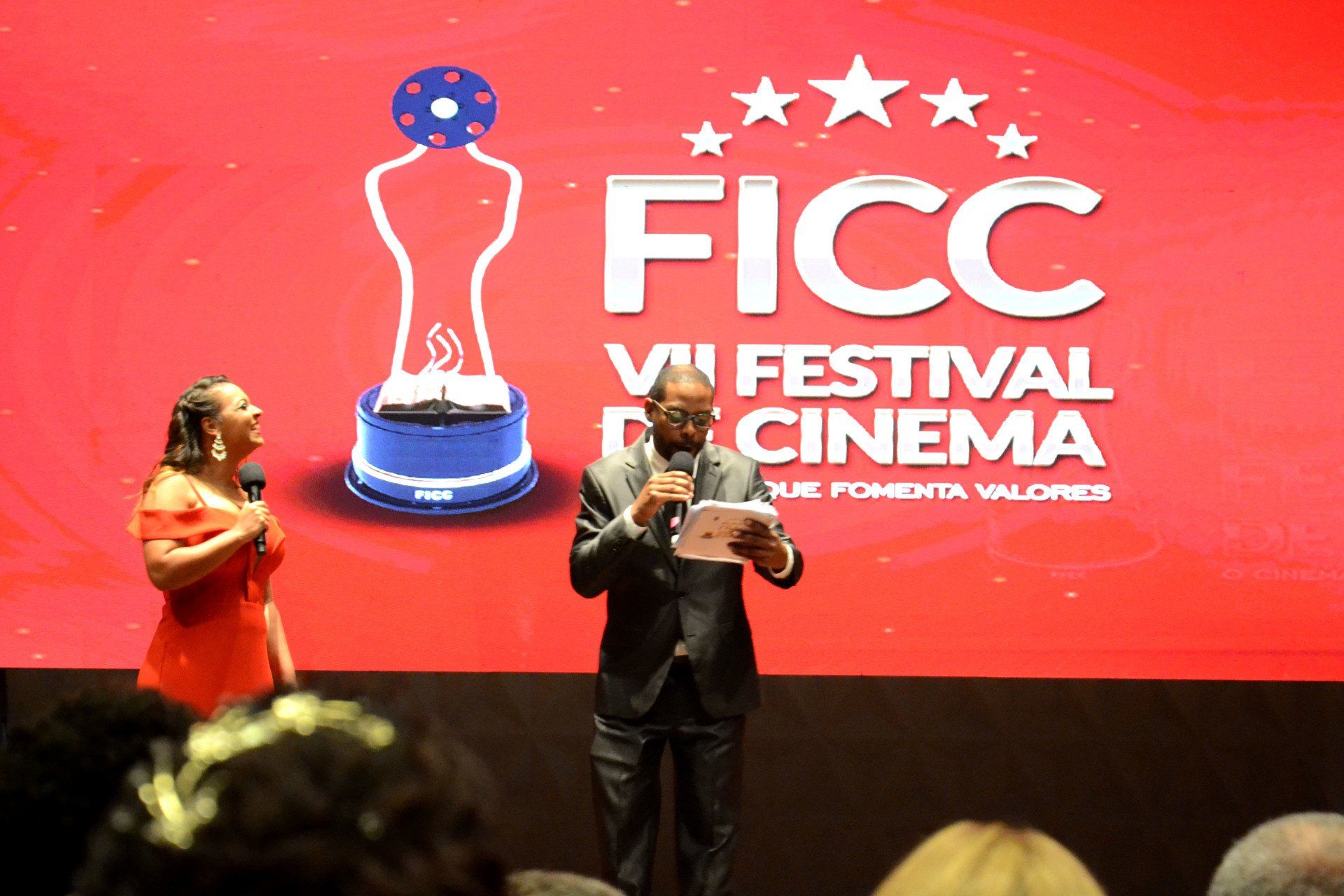 Um homem e uma mulher estão em pé em frente a uma tela que diz cinema festival ficc