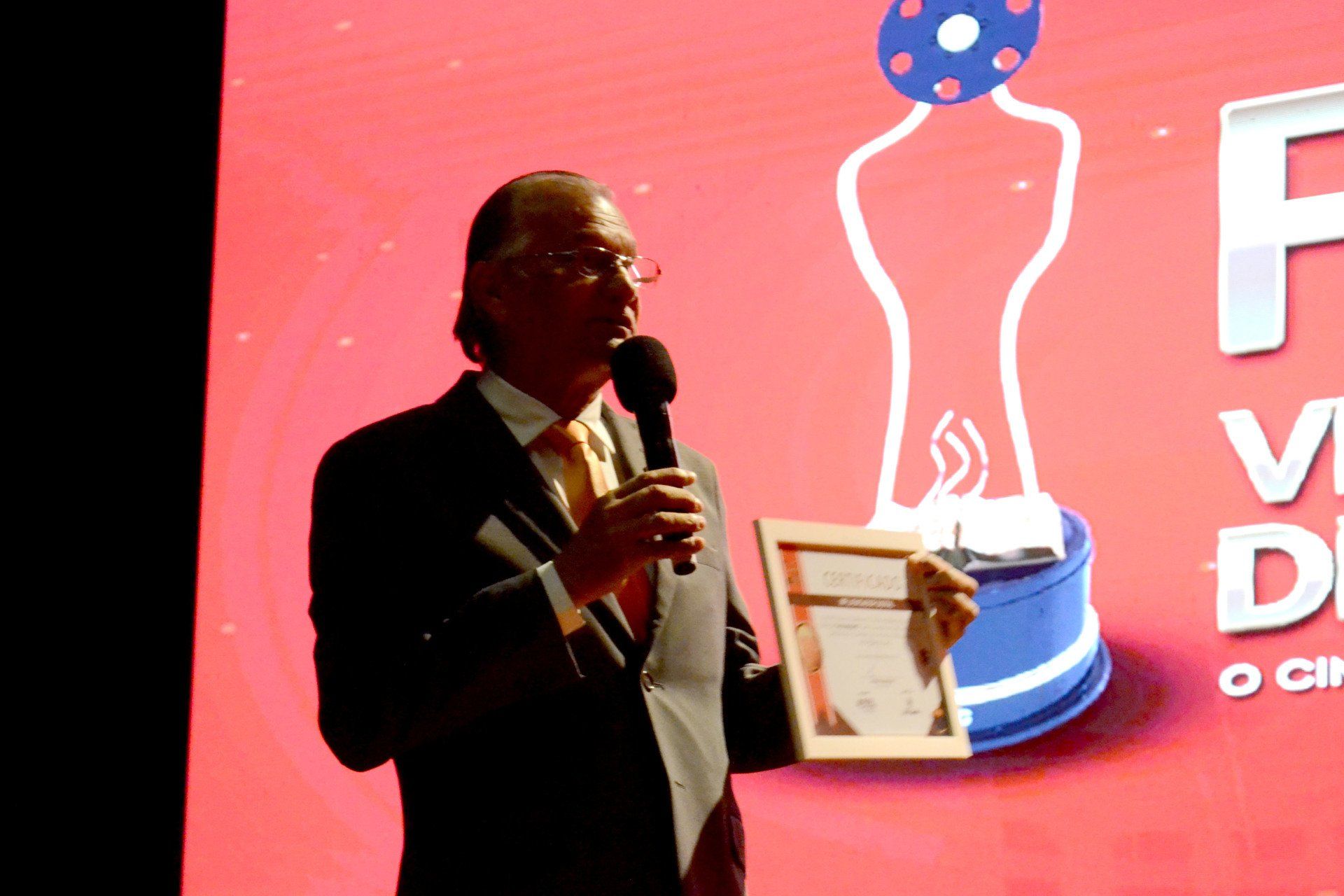Um homem de terno e gravata segura um microfone e um certificado